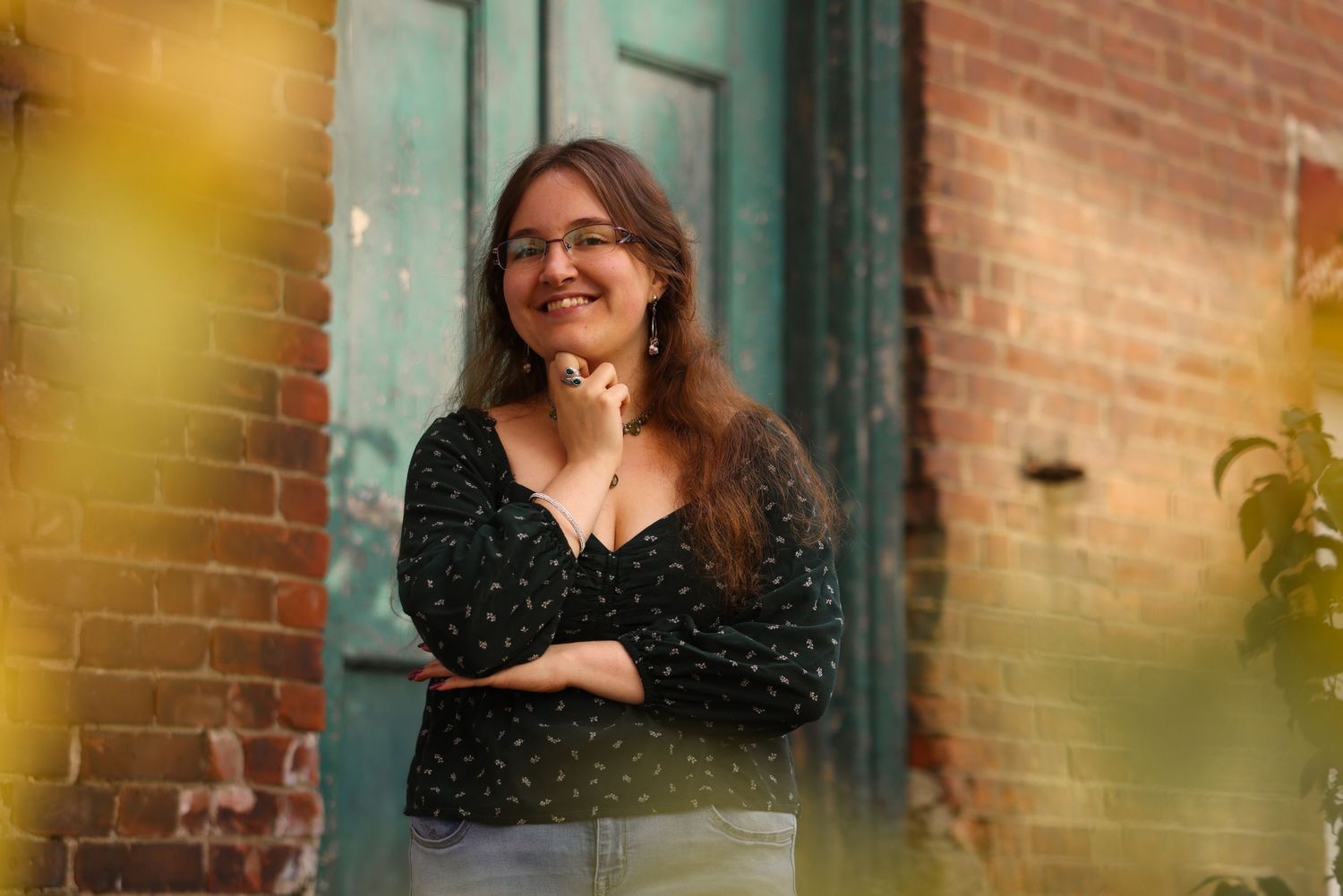 Smiling young woman in black polka dot blouse poses against brick wall with green foliage background.