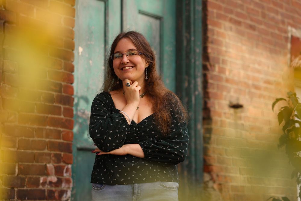 Smiling young woman in black polka dot blouse poses against brick wall with green foliage background.