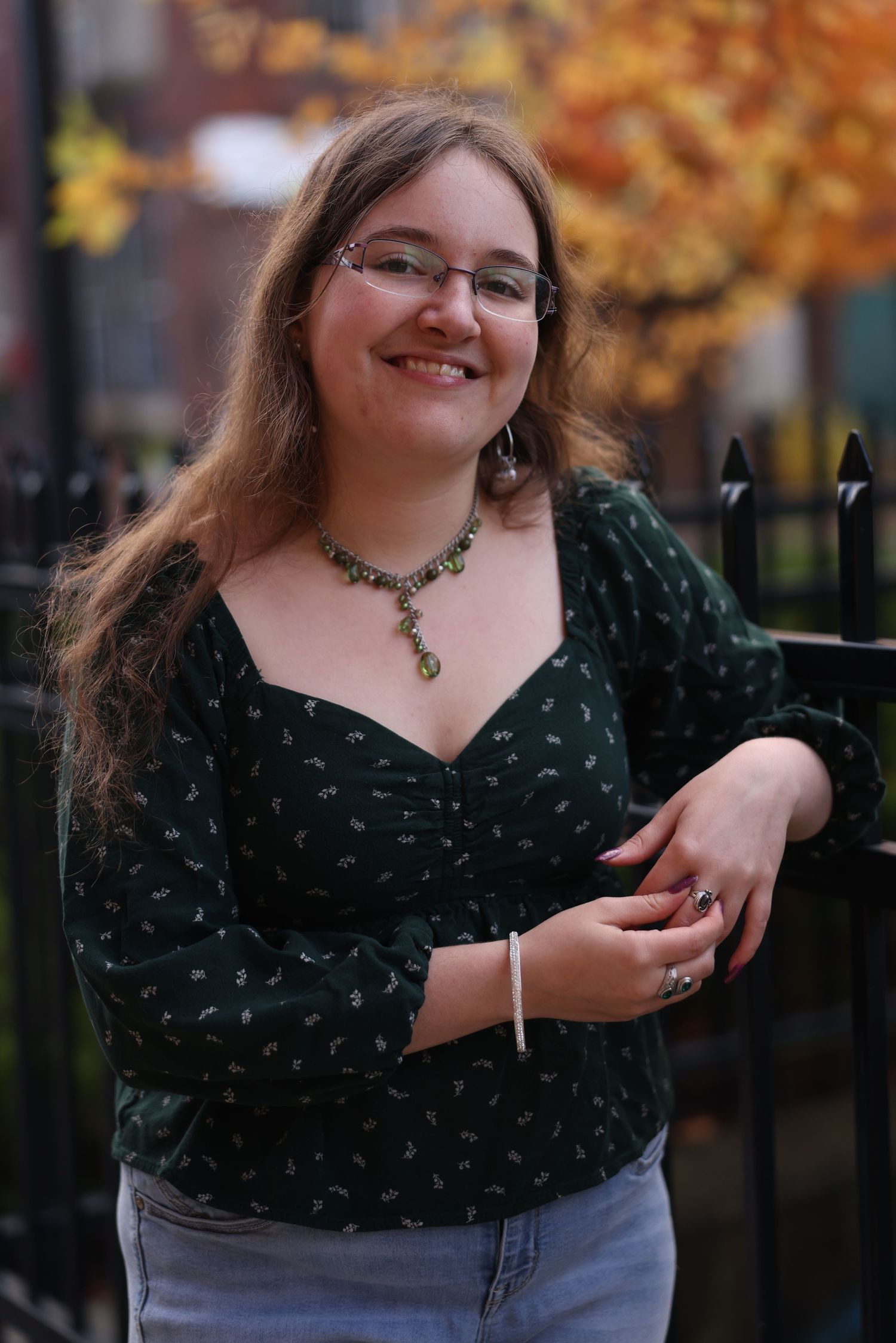 Young woman in green floral top and jeans smiles outdoors near iron fence with autumn foliage background.