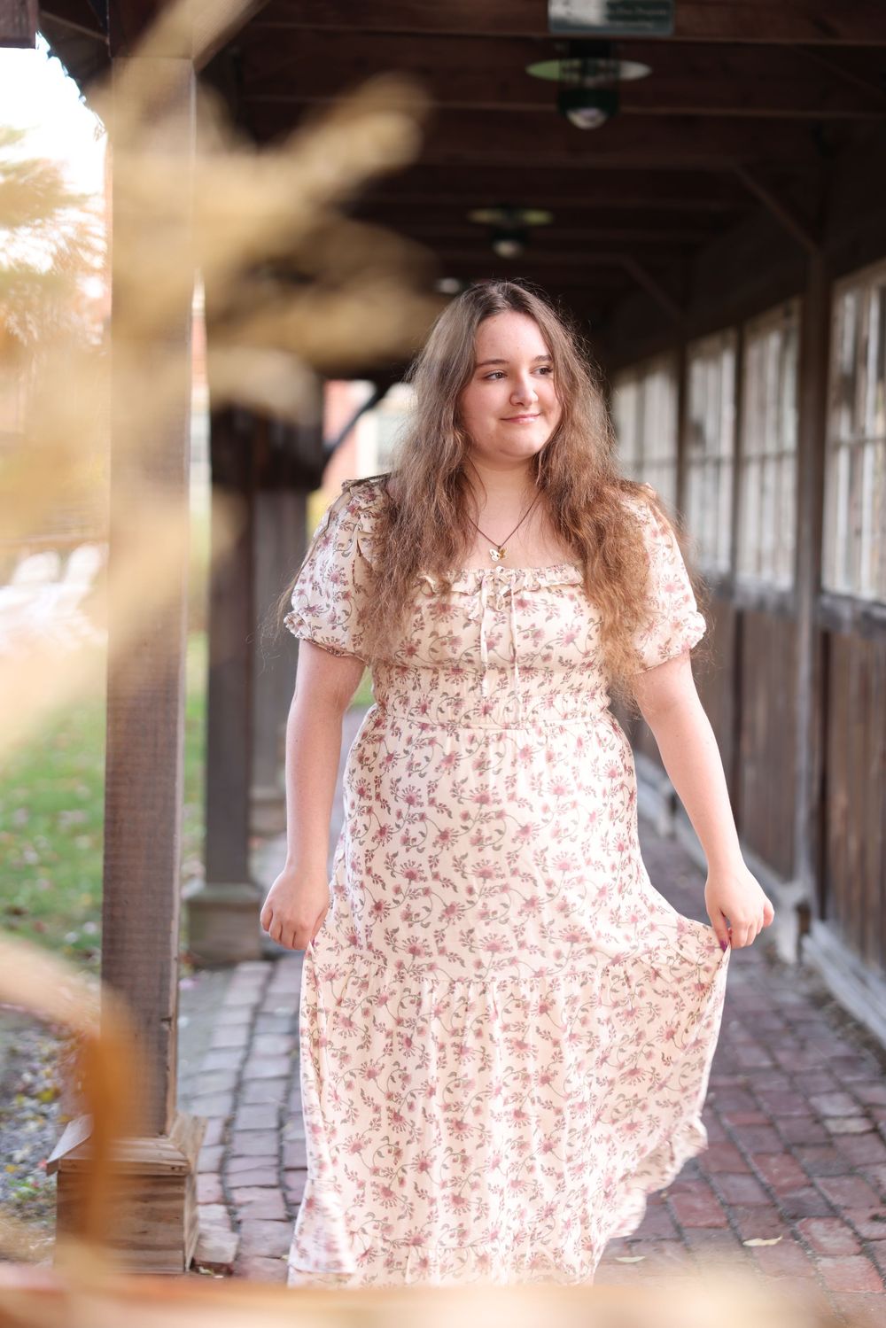 Young woman in floral white maxi dress walks through rustic covered outdoor corridor in warm light.