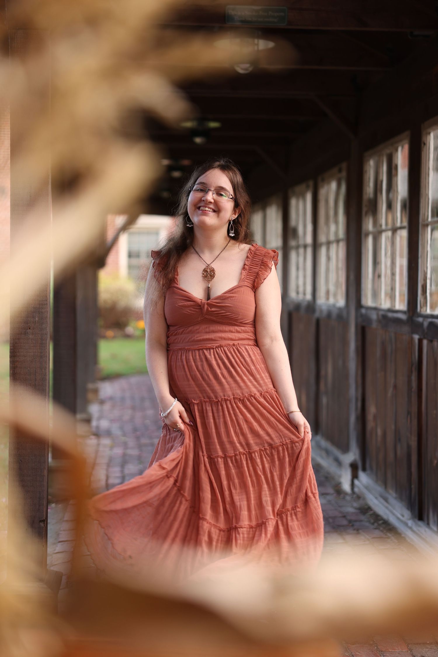 Young woman in rust-colored tiered maxi dress walks through rustic covered walkway in autumn setting.