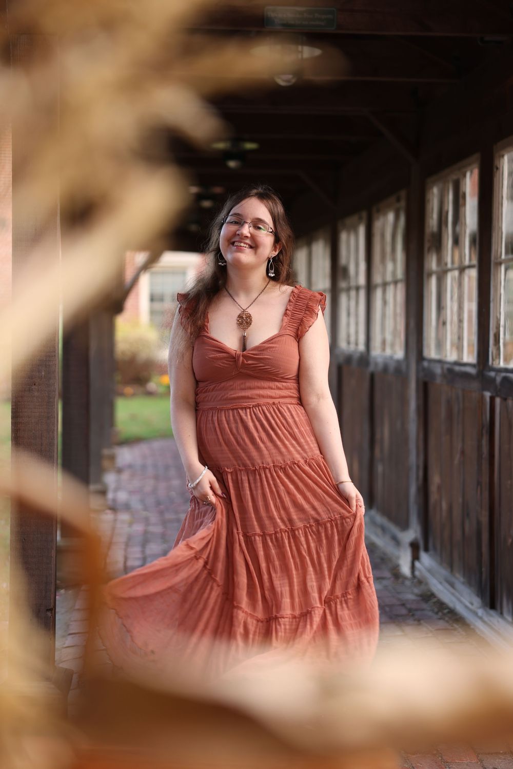 Young woman in rust-colored tiered maxi dress walks through rustic covered walkway in autumn setting.