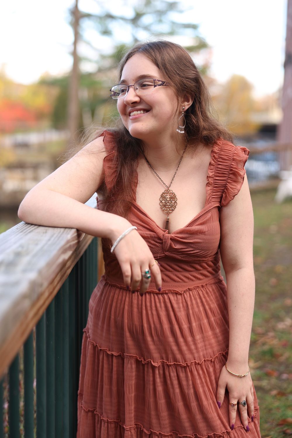 Smiling young woman in rust ruffled dress leans on wooden railing outdoors in fall foliage setting.