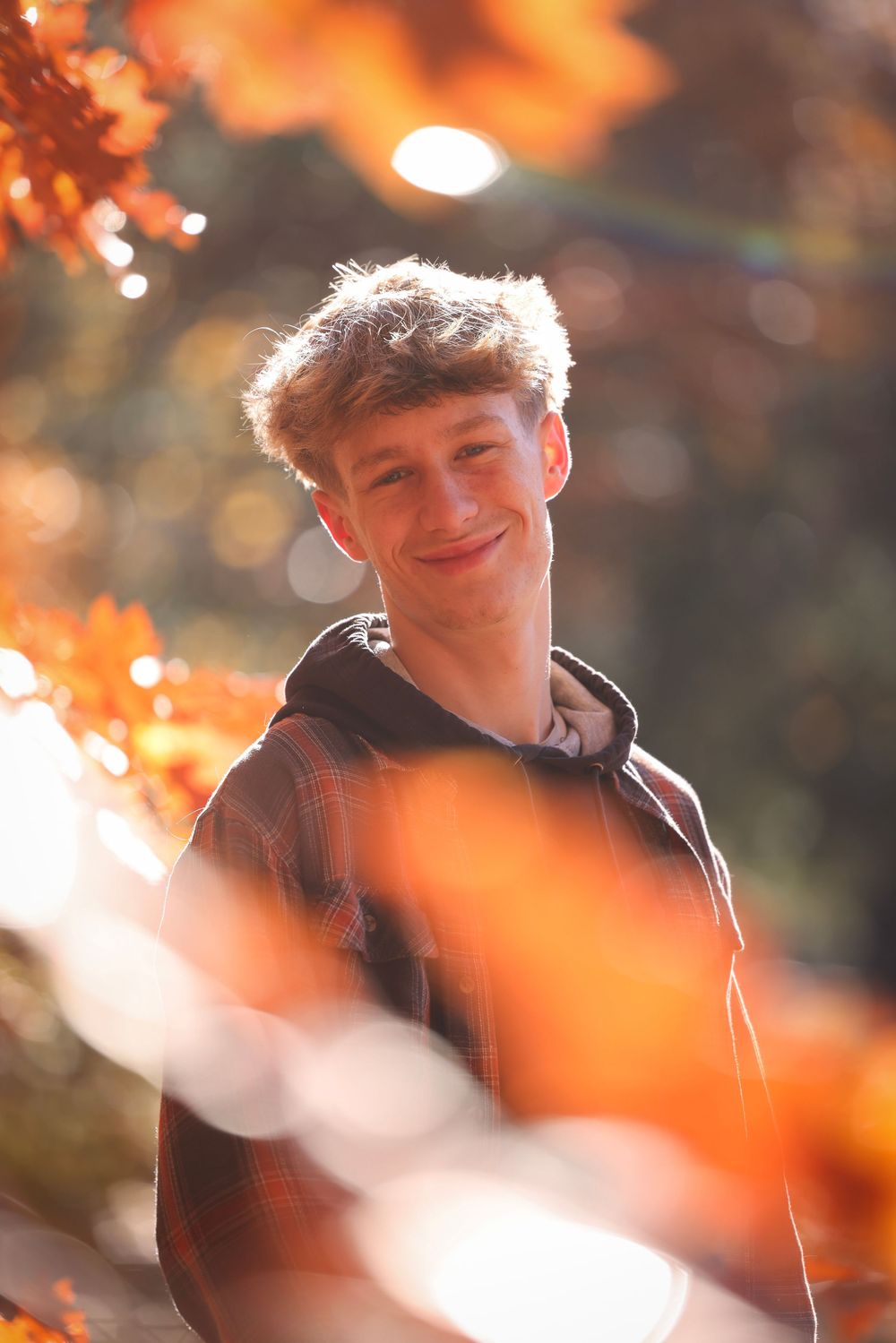 Smiling teenage boy with curly hair poses among glowing orange autumn leaves in warm sunlight.