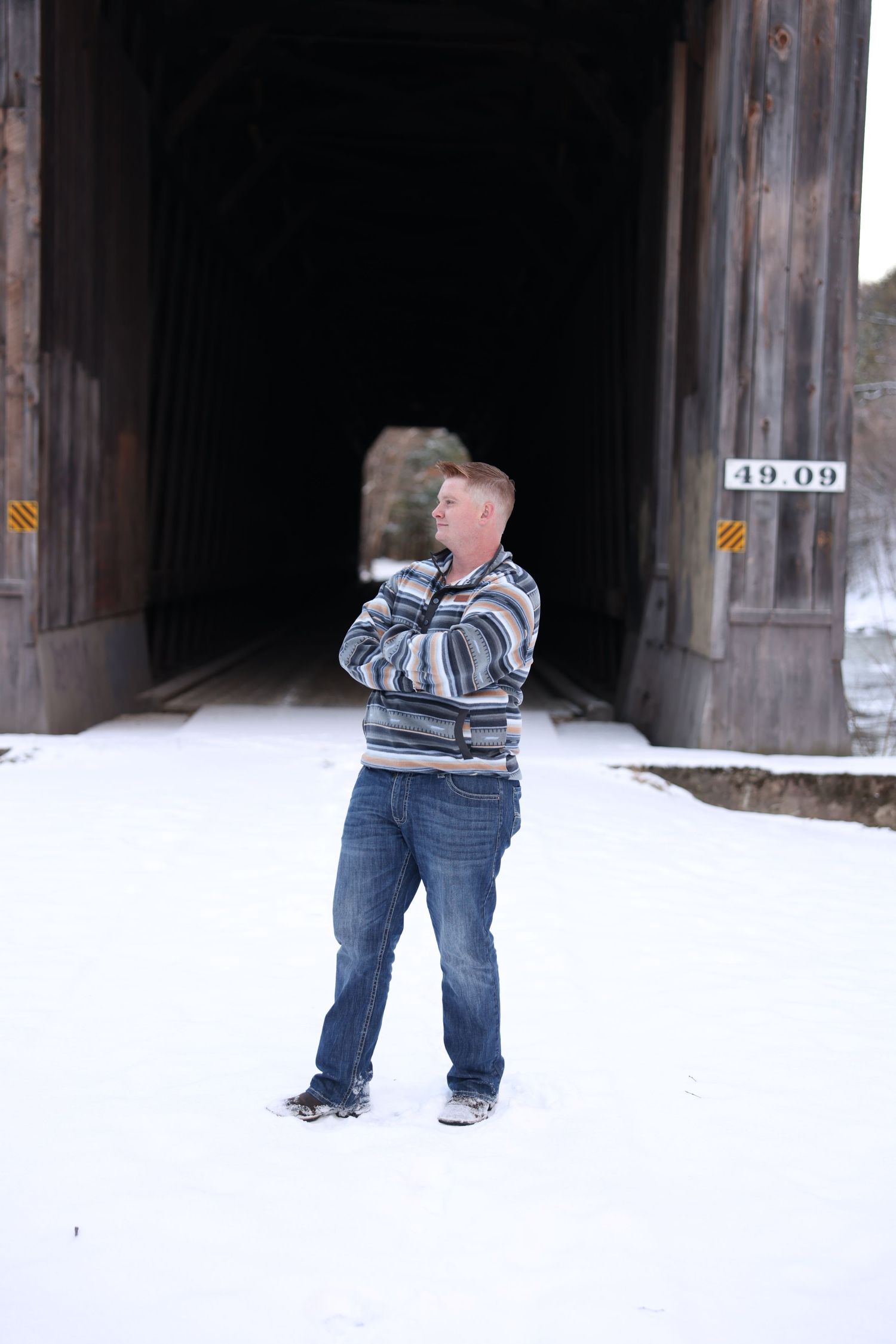 Young man with red hair standing arms crossed in snow before a historic covered wooden bridge.