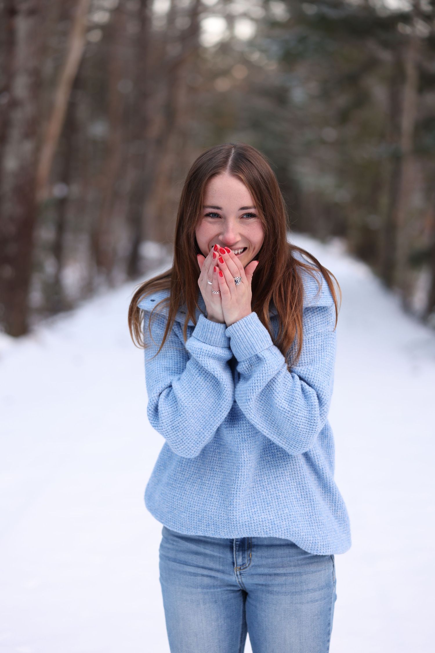 Happy young woman in blue knit sweater covering mouth with red-nailed hands in snowy winter forest.