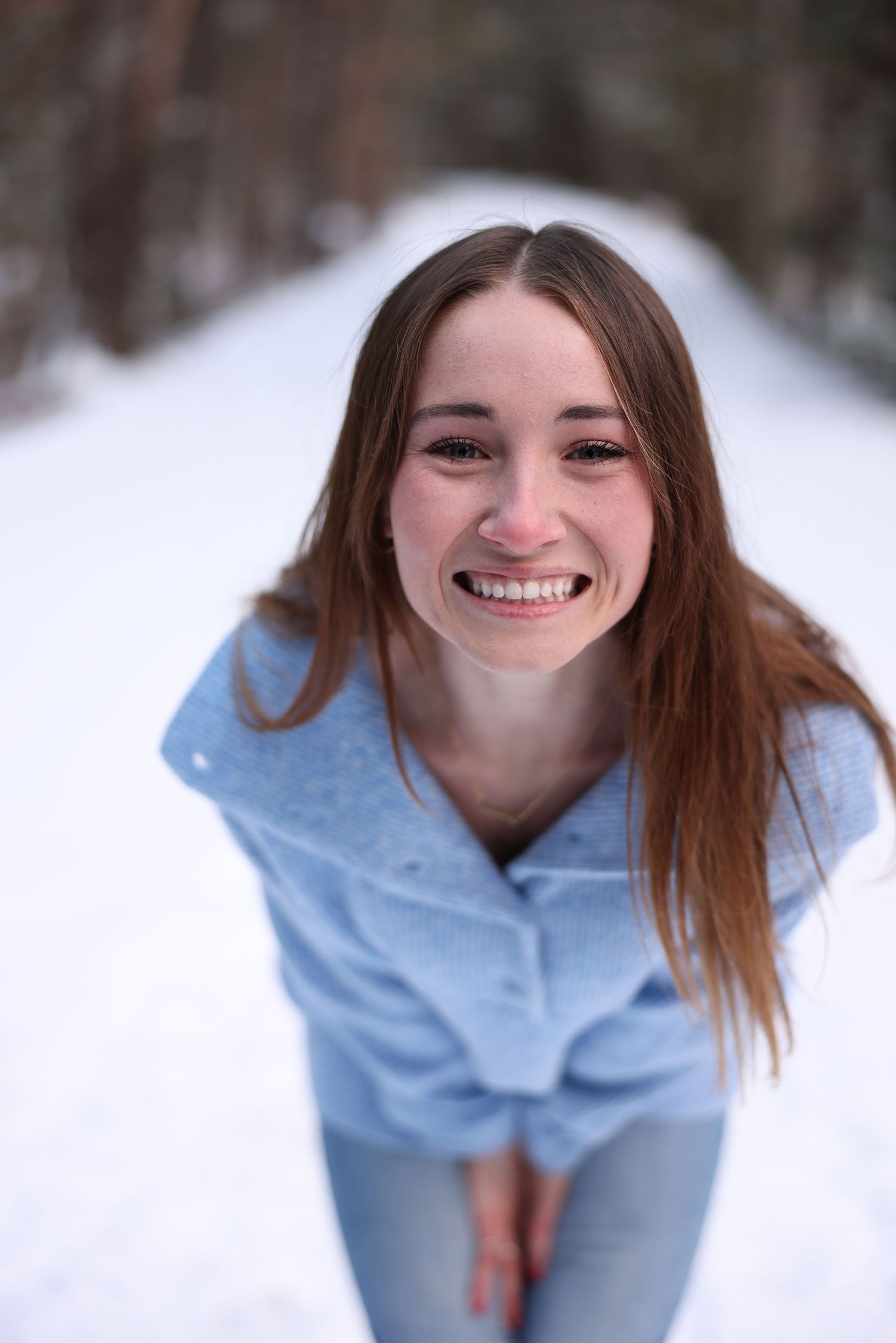 Cheerful brunette teen girl in blue sweater smiling on a snowy forest path in winter.