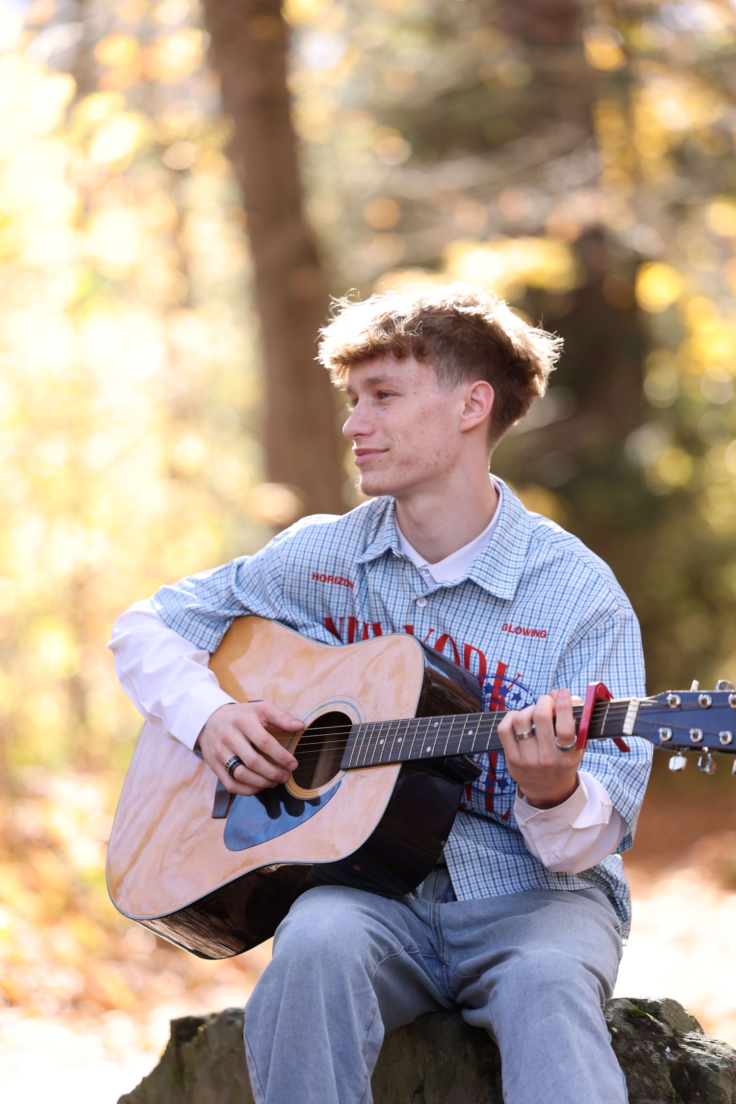Teen boy plays acoustic guitar seated on a rock in a bright autumn forest setting.