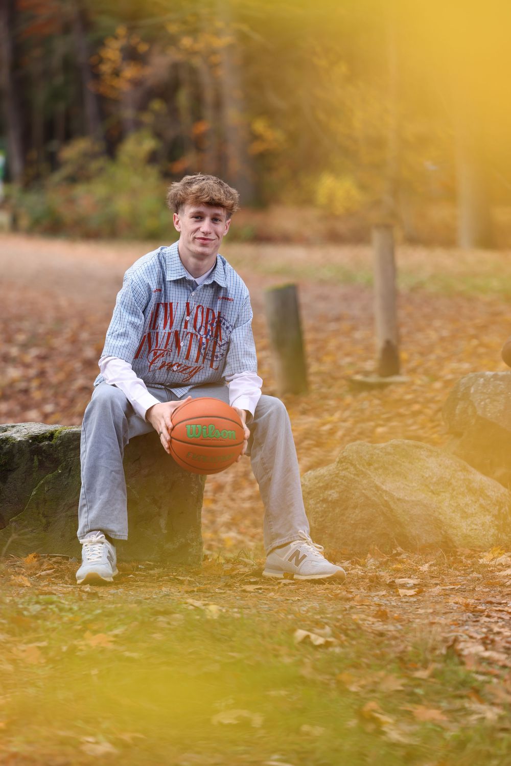 Teen boy holds basketball while seated on a rock in a golden autumn park setting.