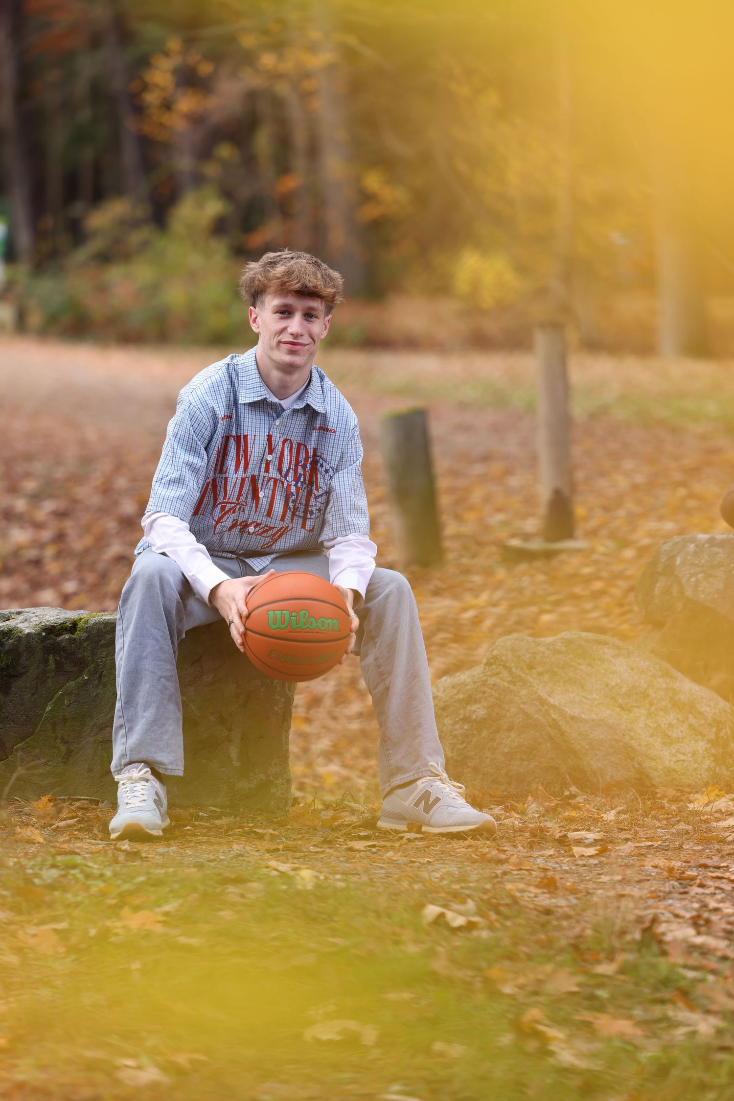 Teen boy holds basketball while seated on a rock in a golden autumn park setting.
