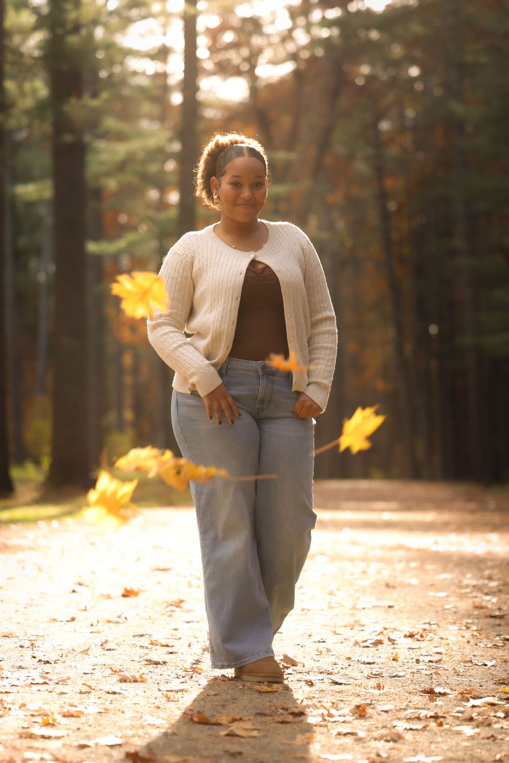 Young Black woman in cream cardigan and jeans walks forest path surrounded by golden fall foliage.