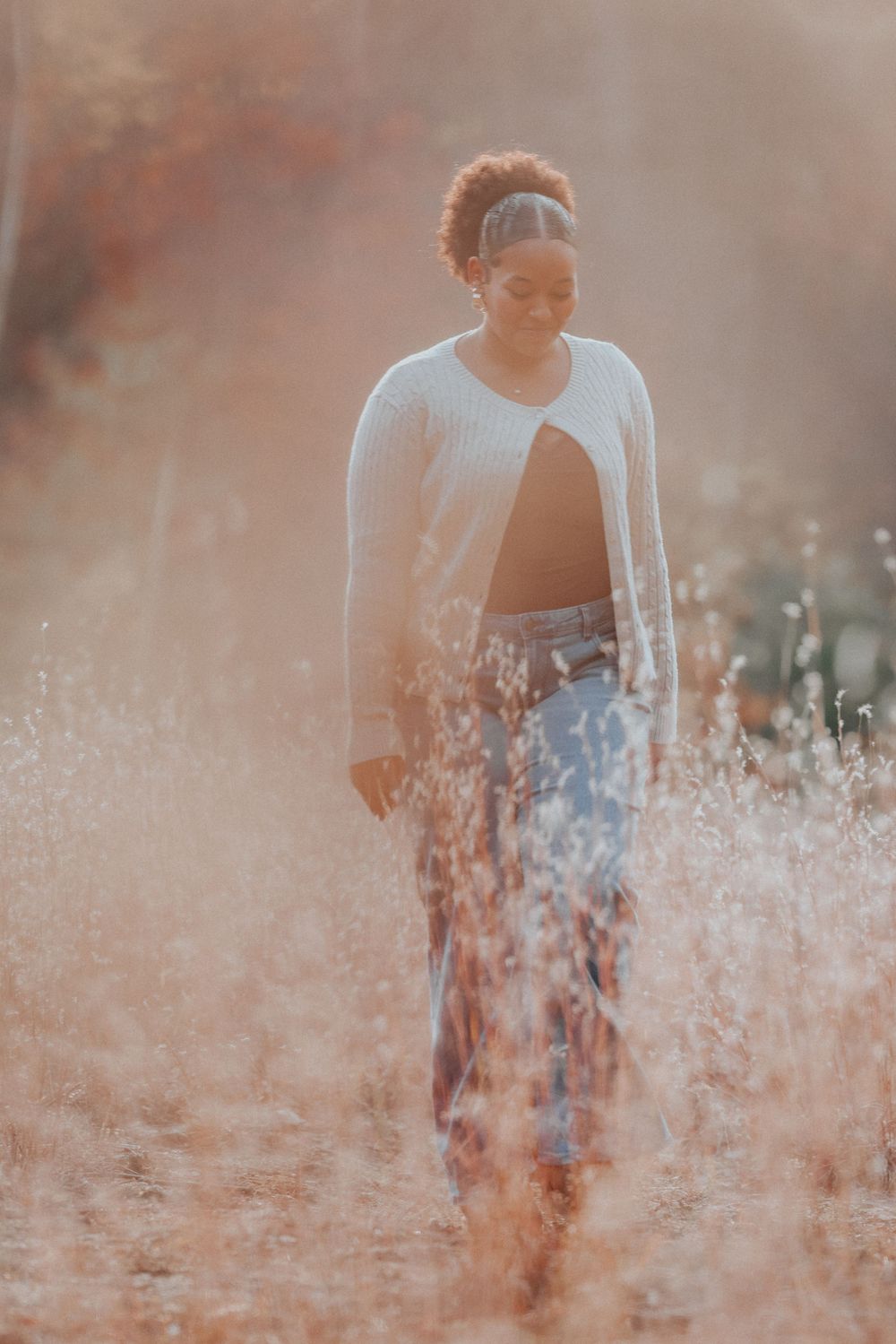 A young Black woman walks through golden dry grass fields wearing a white cardigan and floral pants in warm autumn light.