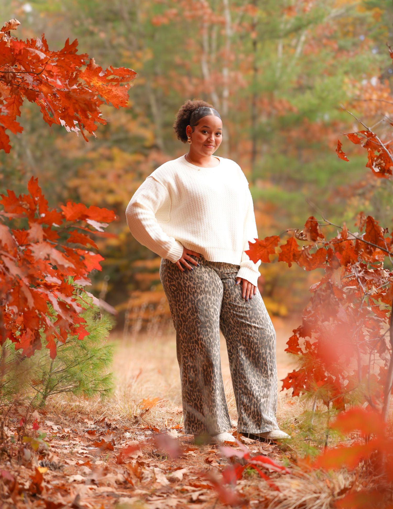 Young Black woman in white sweater and leopard print pants poses among vibrant red autumn leaves.