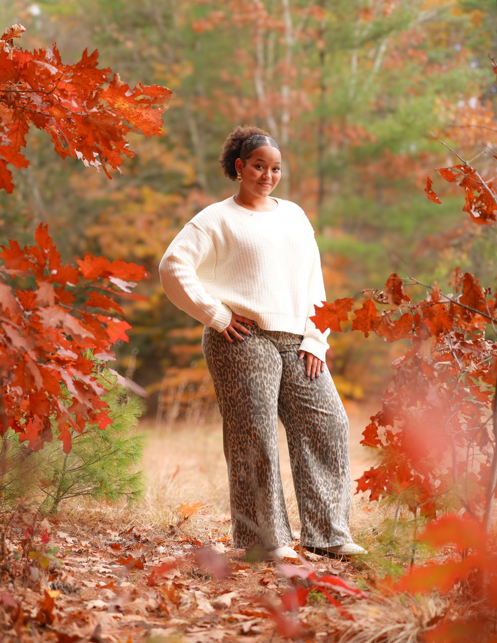 Young Black woman in white sweater and leopard print pants poses among vibrant red autumn leaves.