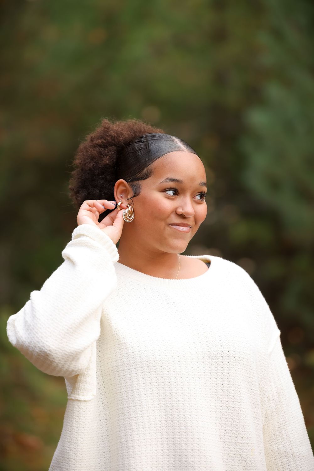 Young Black woman in white sweater touches her gold earring outdoors with green foliage background.