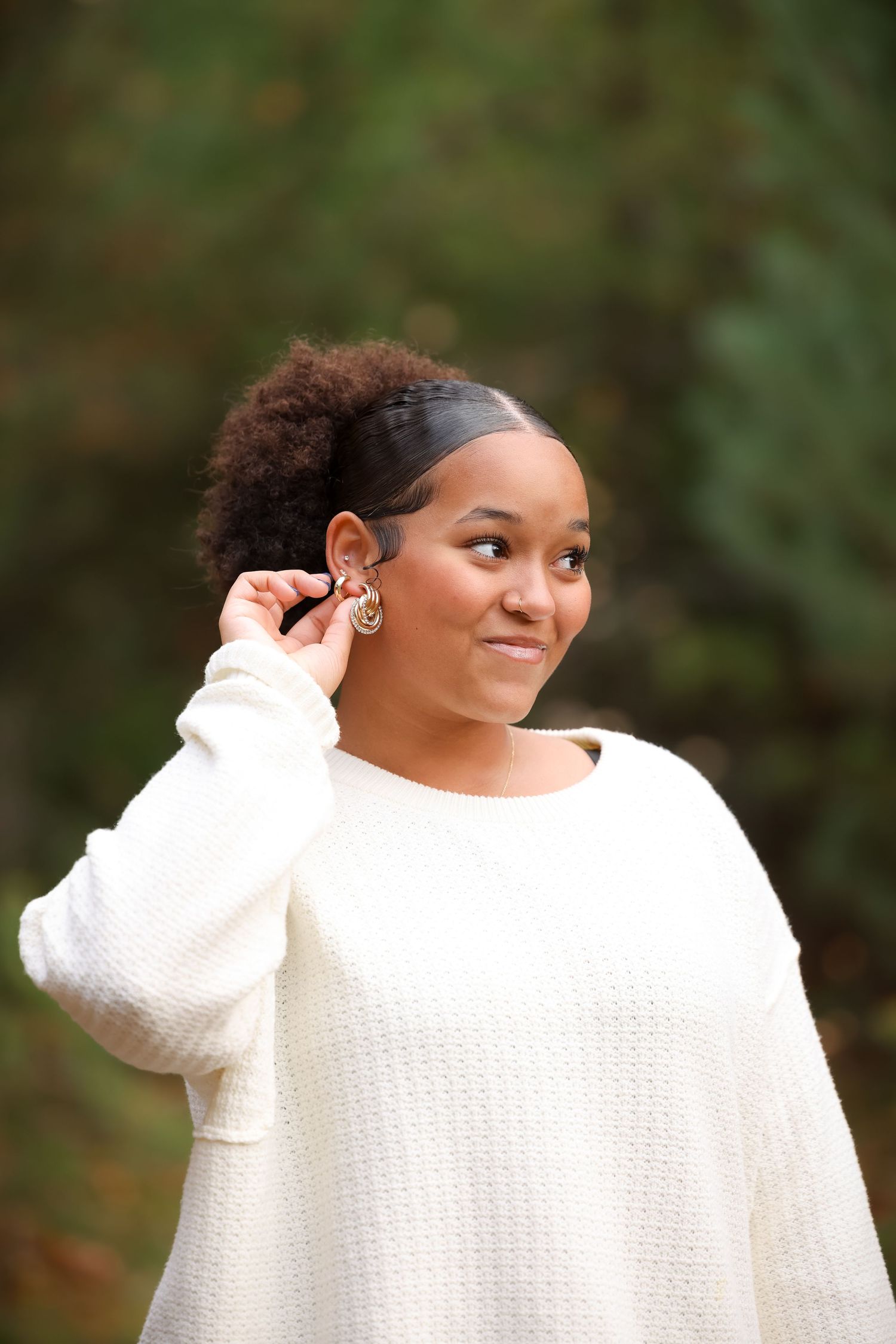Young Black woman in white sweater touches her gold earring outdoors with green foliage background.