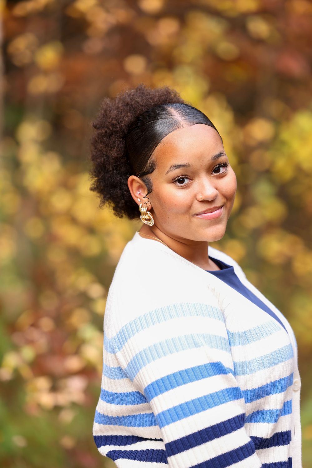 Young woman in striped cardigan smiling outdoors with colorful fall foliage background.