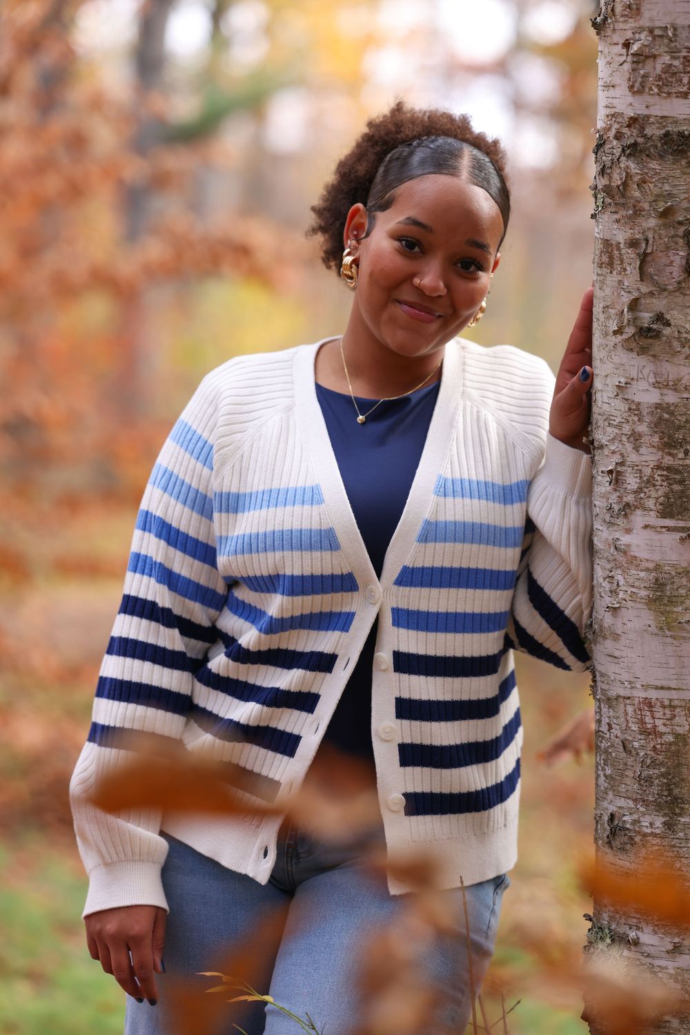Young woman in striped cardigan leaning against a birch tree in colorful autumn foliage.