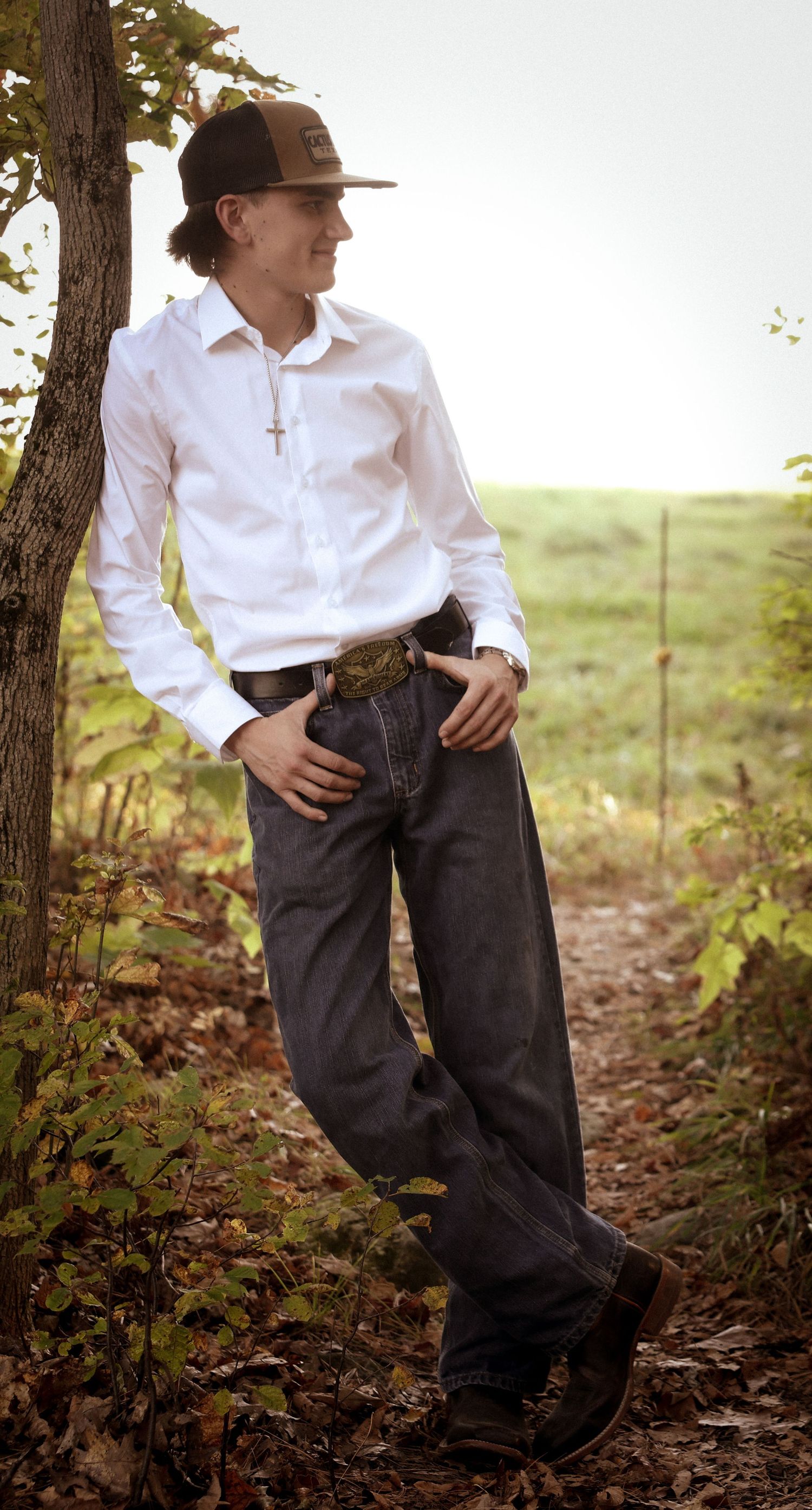 A young man in a white shirt, dark jeans, and Cactus Ropes cap leans against a tree on a woodland path.