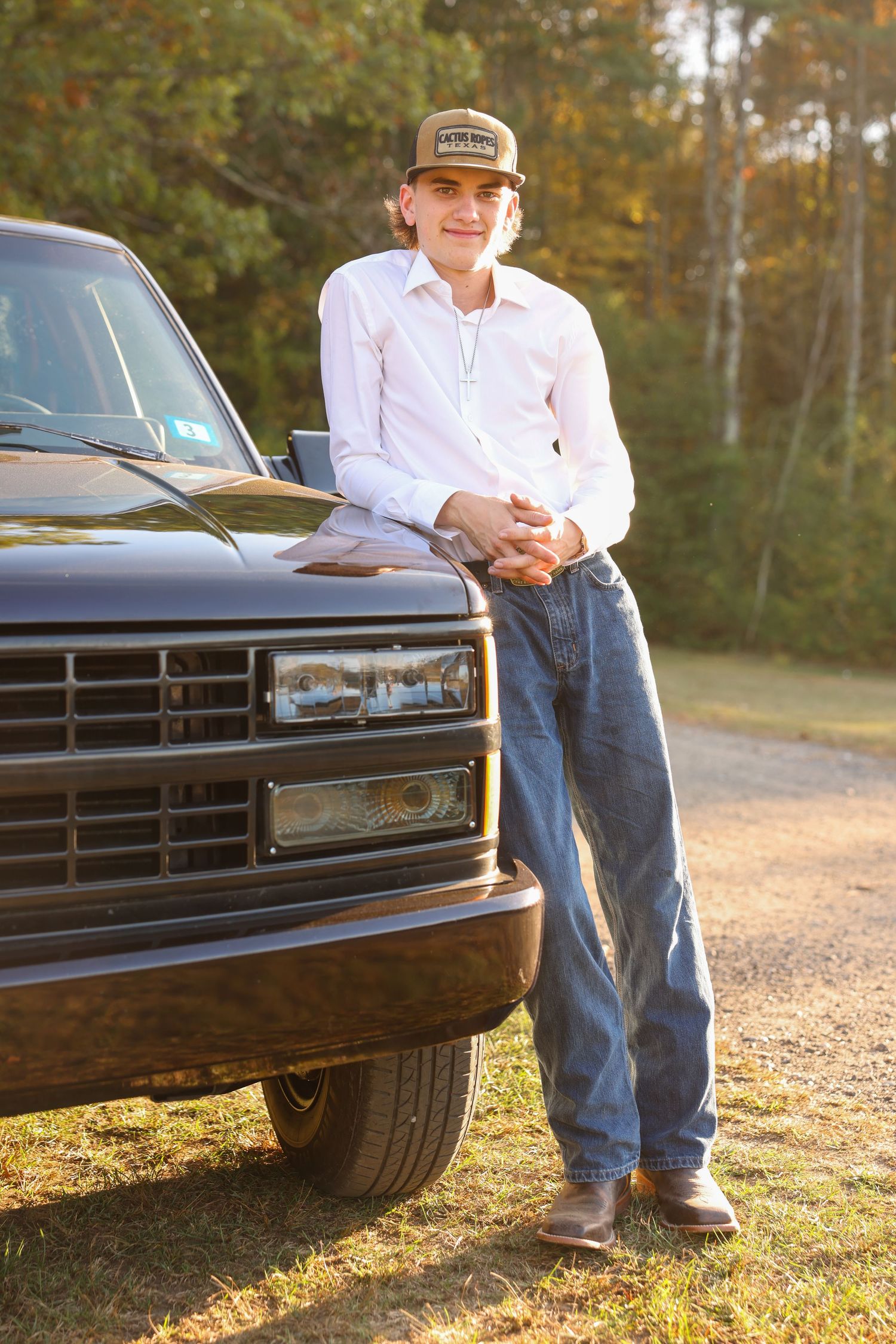 A smiling young man in a white shirt and jeans leans against a classic black truck in autumn sunlight.