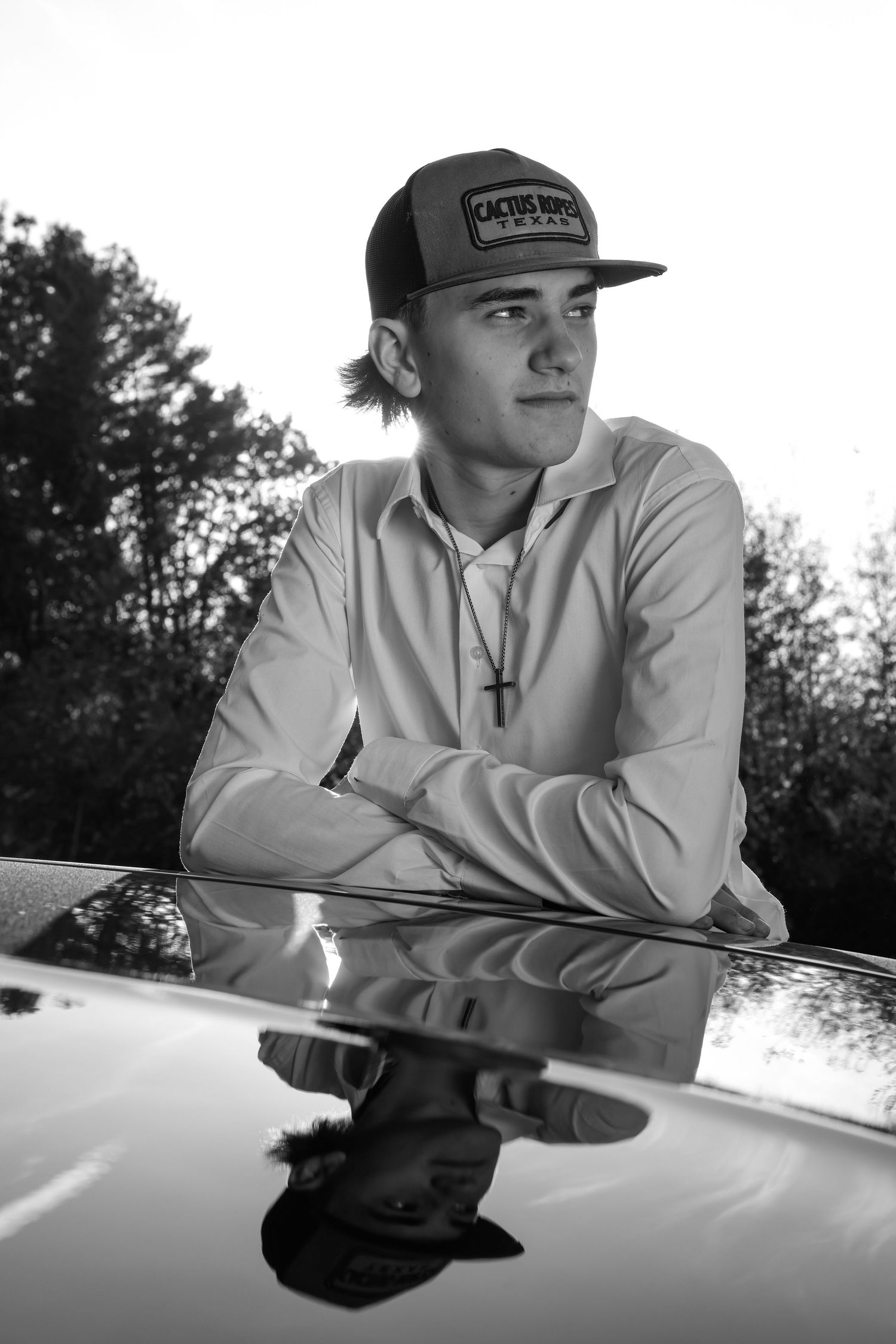 Black and white portrait of a young man in a cap leaning on a car hood with his reflection visible.