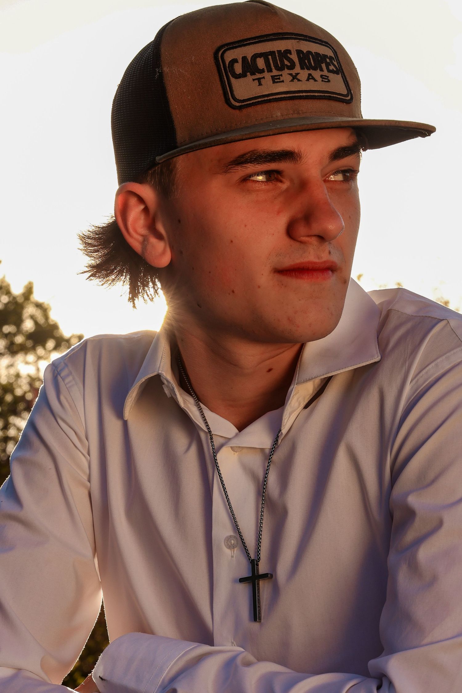 Close-up portrait of a young man wearing a Cactus Ropes Texas cap and cross necklace in golden backlight.