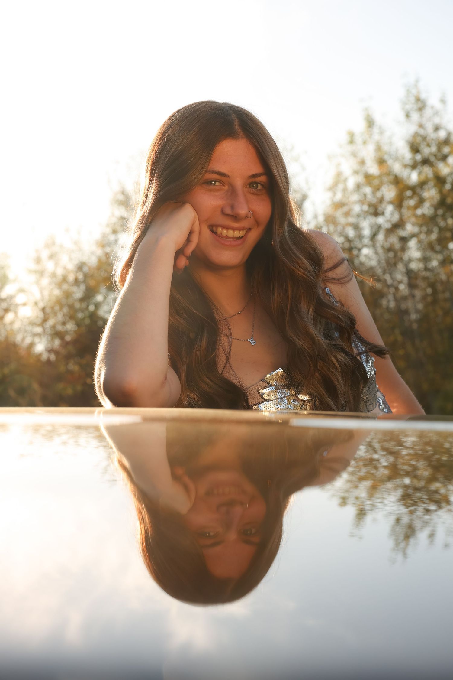 A smiling young woman with long wavy hair leans on a reflective car roof in warm golden sunset light.