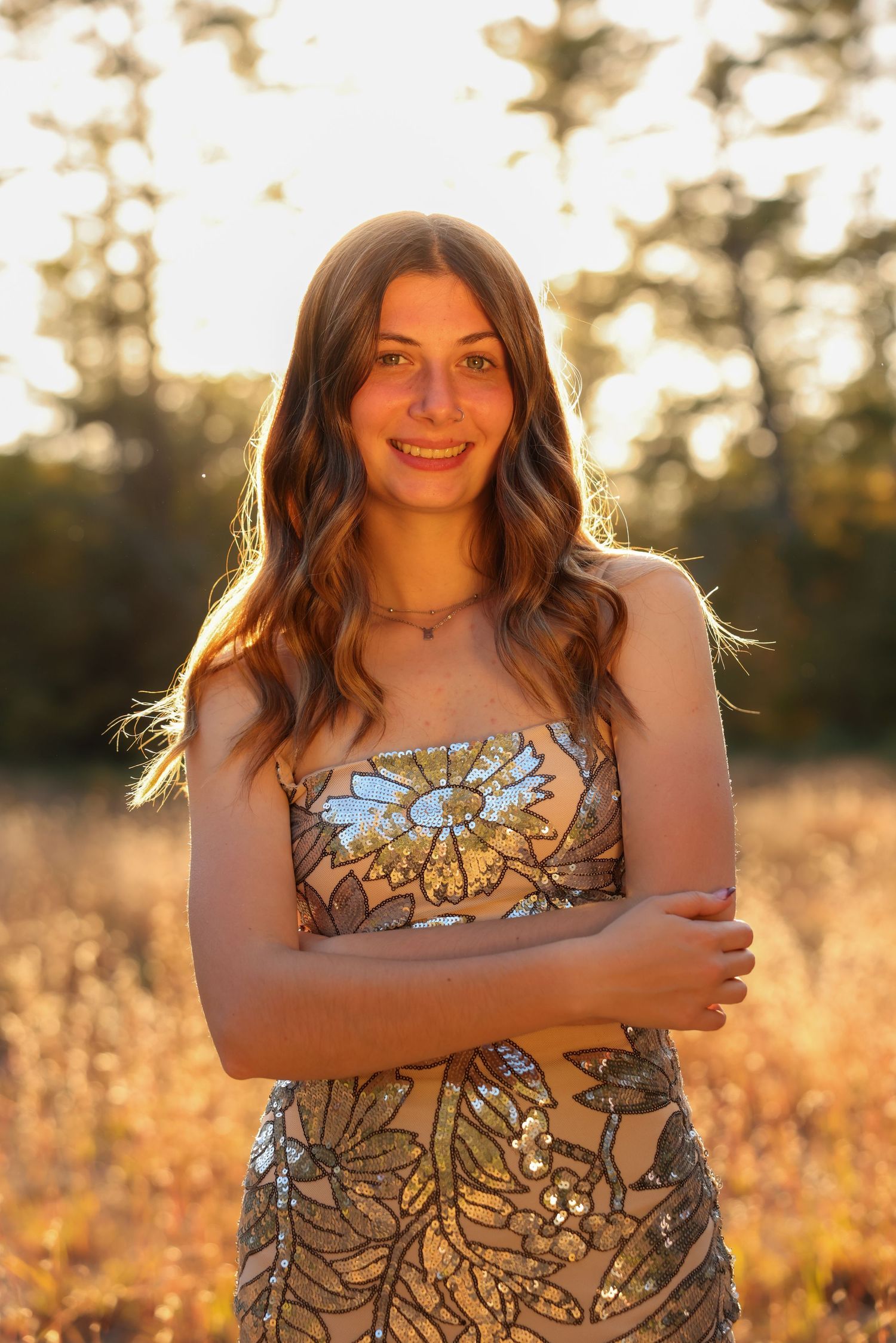 A smiling young woman in a strapless sequined dress stands with arms crossed in a golden sunlit meadow.