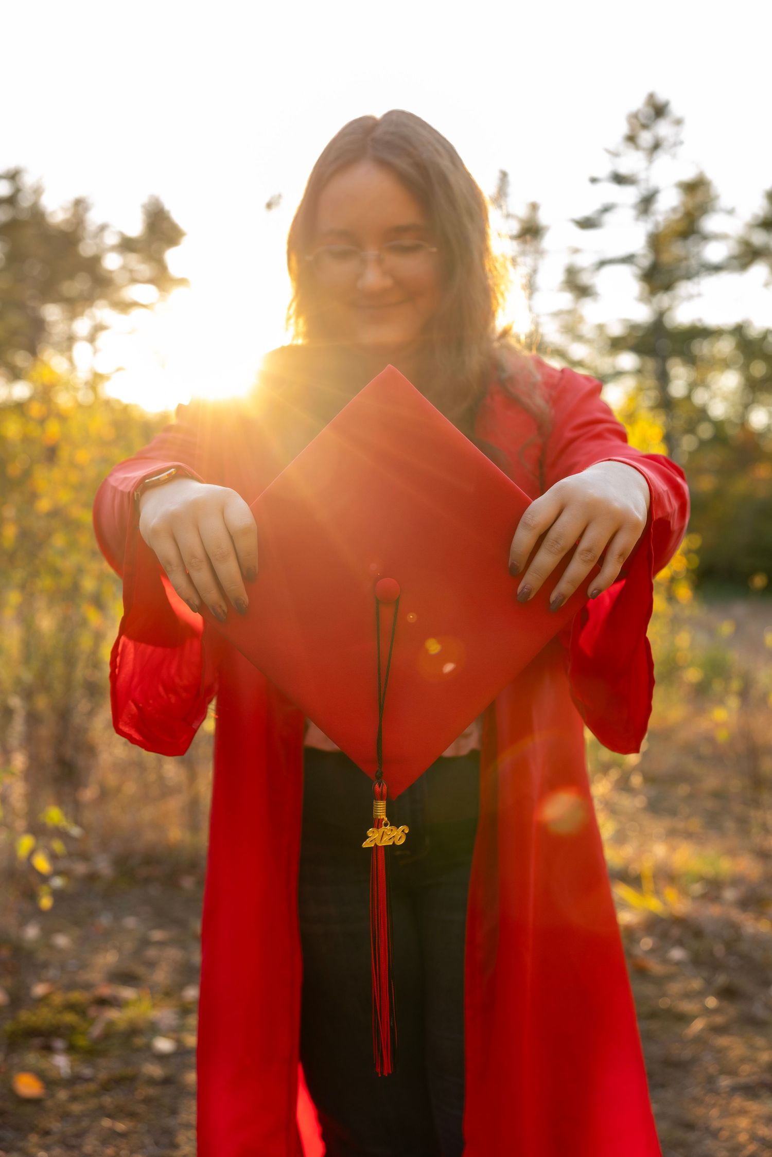 Graduate in red cap and gown holds mortarboard toward camera in glowing sunset outdoor setting.
