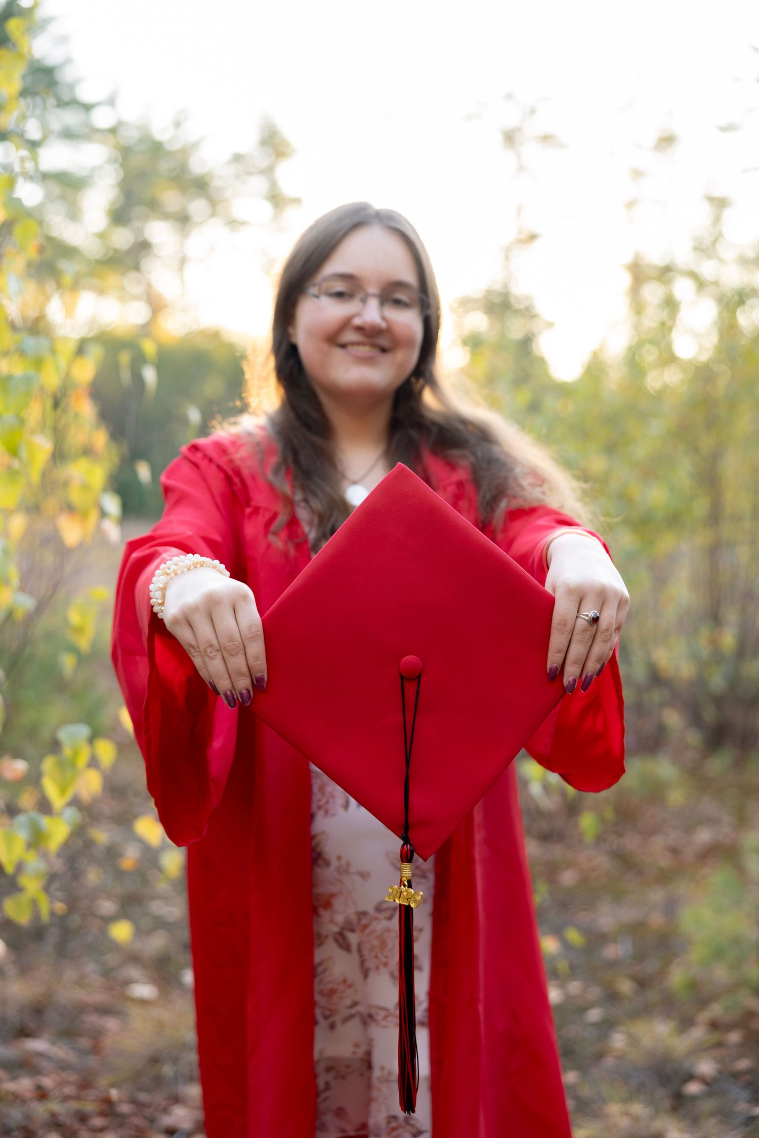 Smiling graduate in red cap and gown holds mortarboard forward in an outdoor autumn setting.