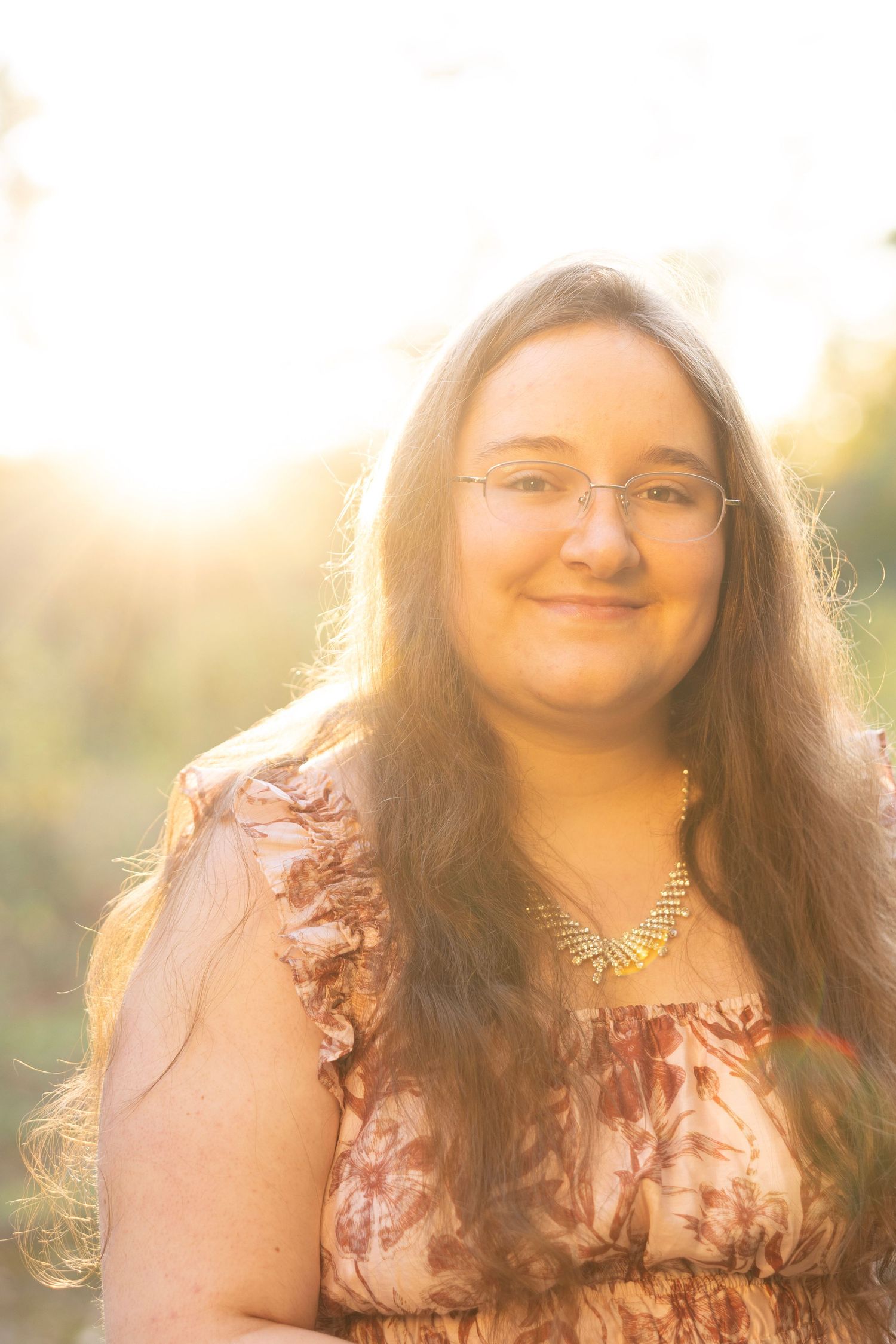 Smiling young woman with long brown hair and glasses wears floral dress in golden sunlight outdoors.