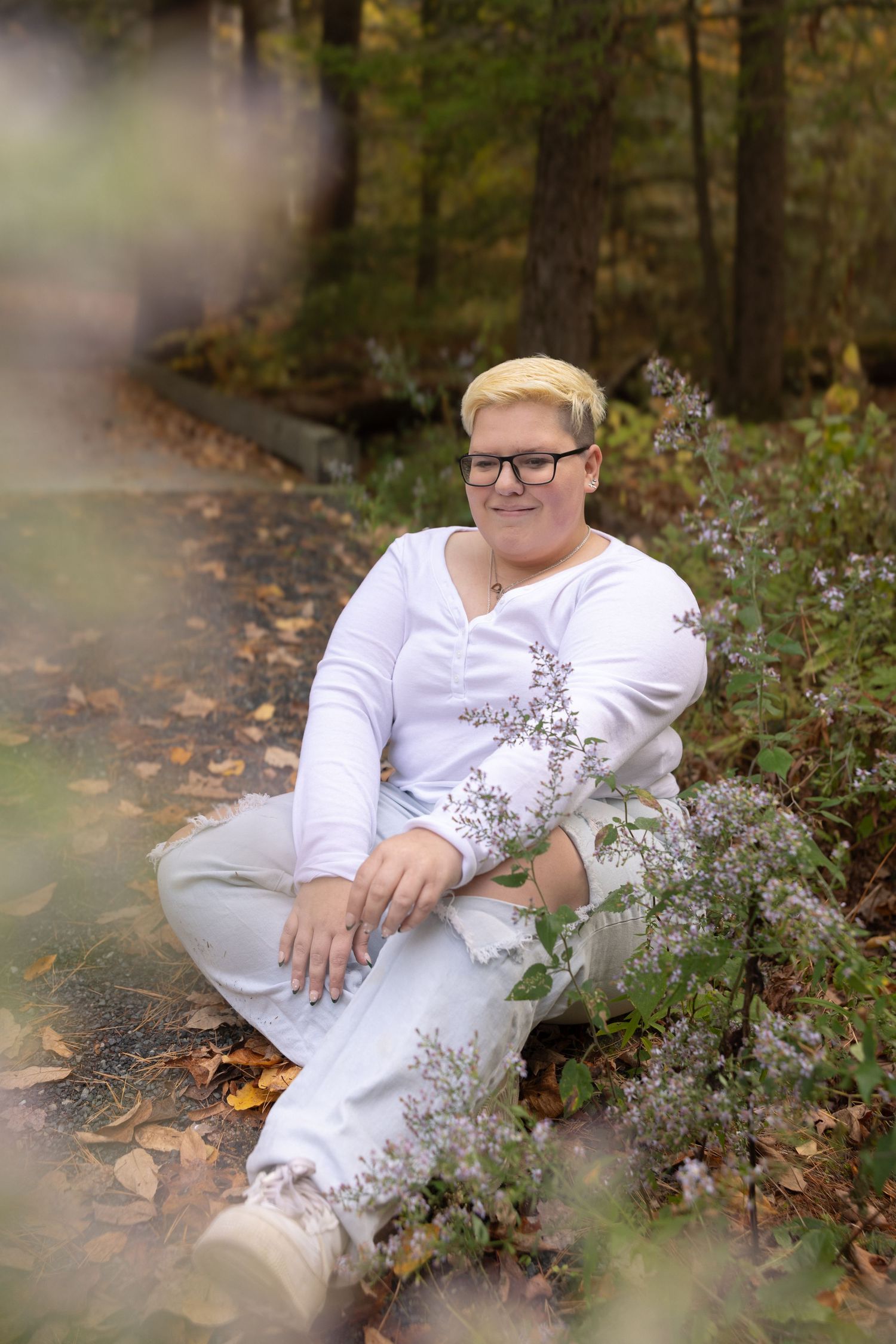 Woman with short blonde hair in white outfit sitting among wildflowers in autumn forest.