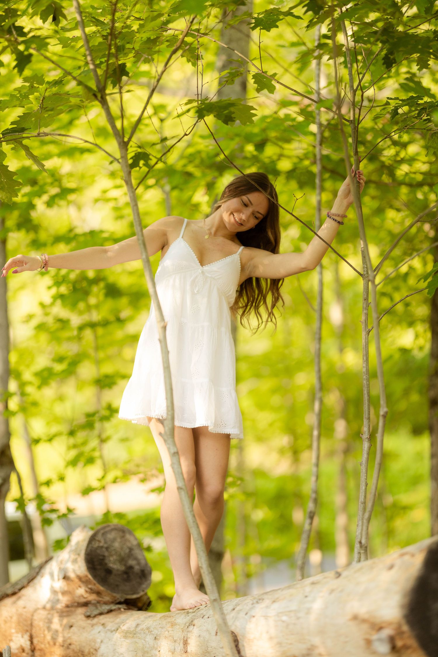 Young woman in white sundress balancing on a fallen log among green trees in a sunlit forest.