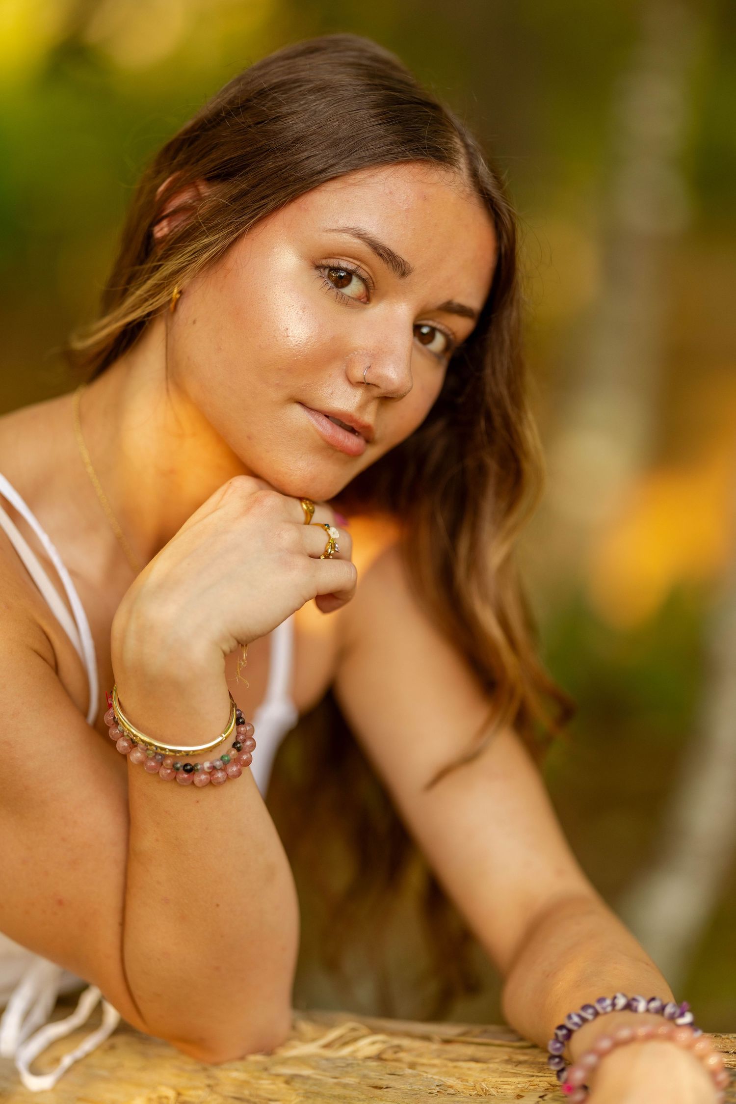 Young woman with long brown hair wearing gold bracelets and white top poses outdoors in warm golden light.