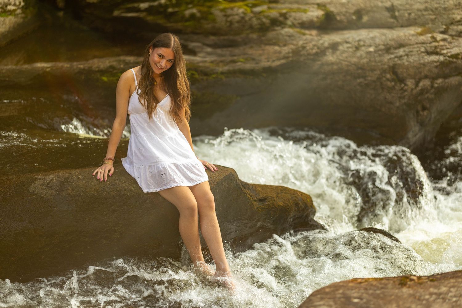 Young woman in white dress sitting on rocks beside rushing river rapids in a sunlit forest setting.
