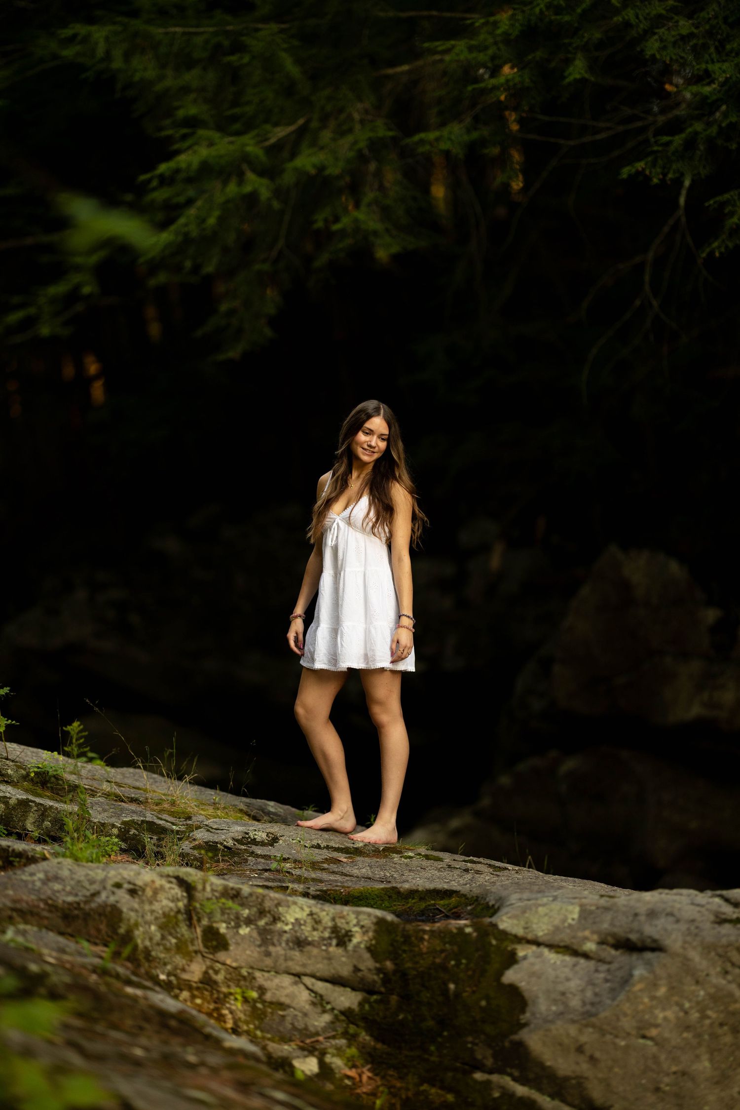 Young woman in white dress standing barefoot on rocks in a dark forest setting.