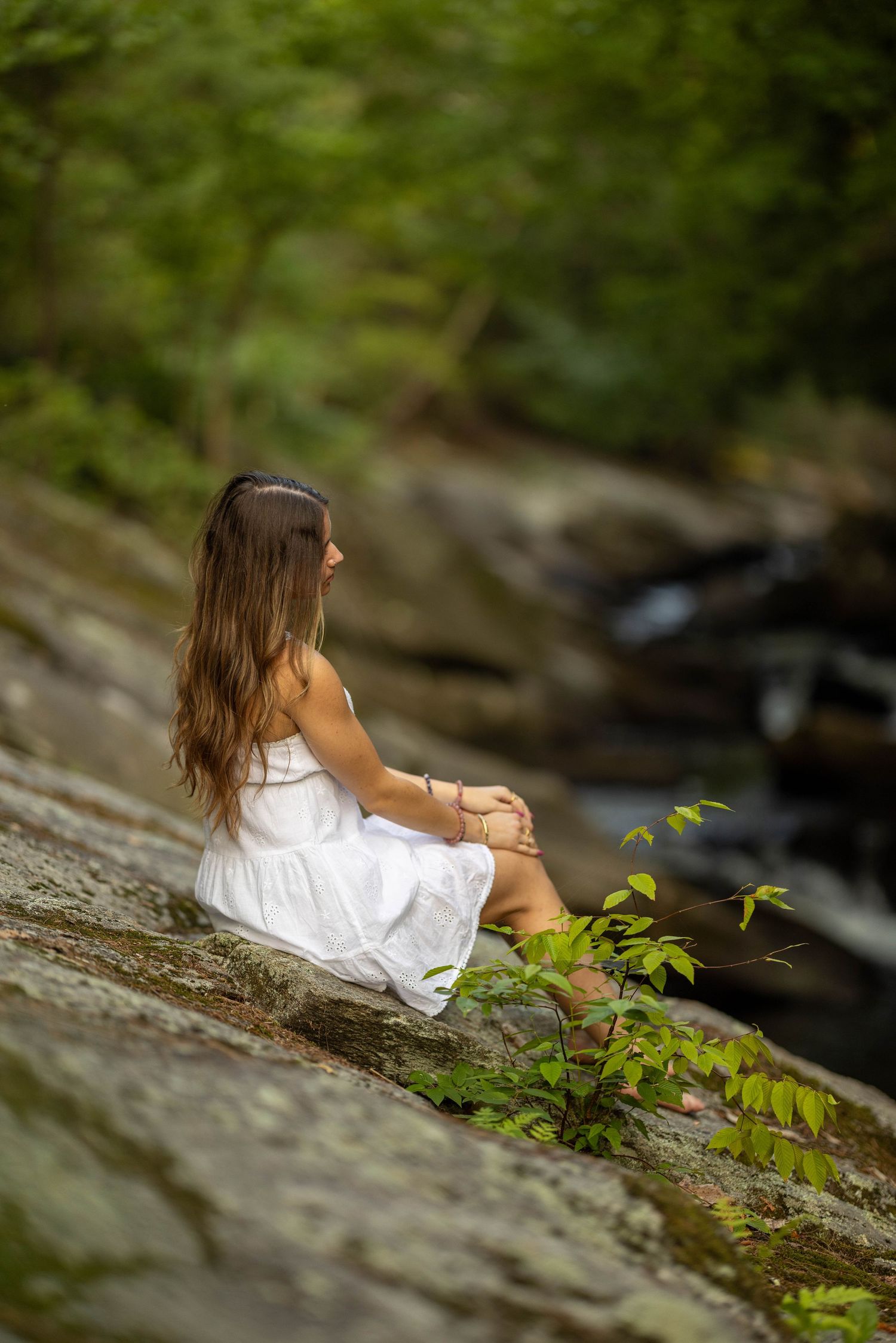Young woman in white dress sitting peacefully on rocks beside a forest stream surrounded by lush green trees.