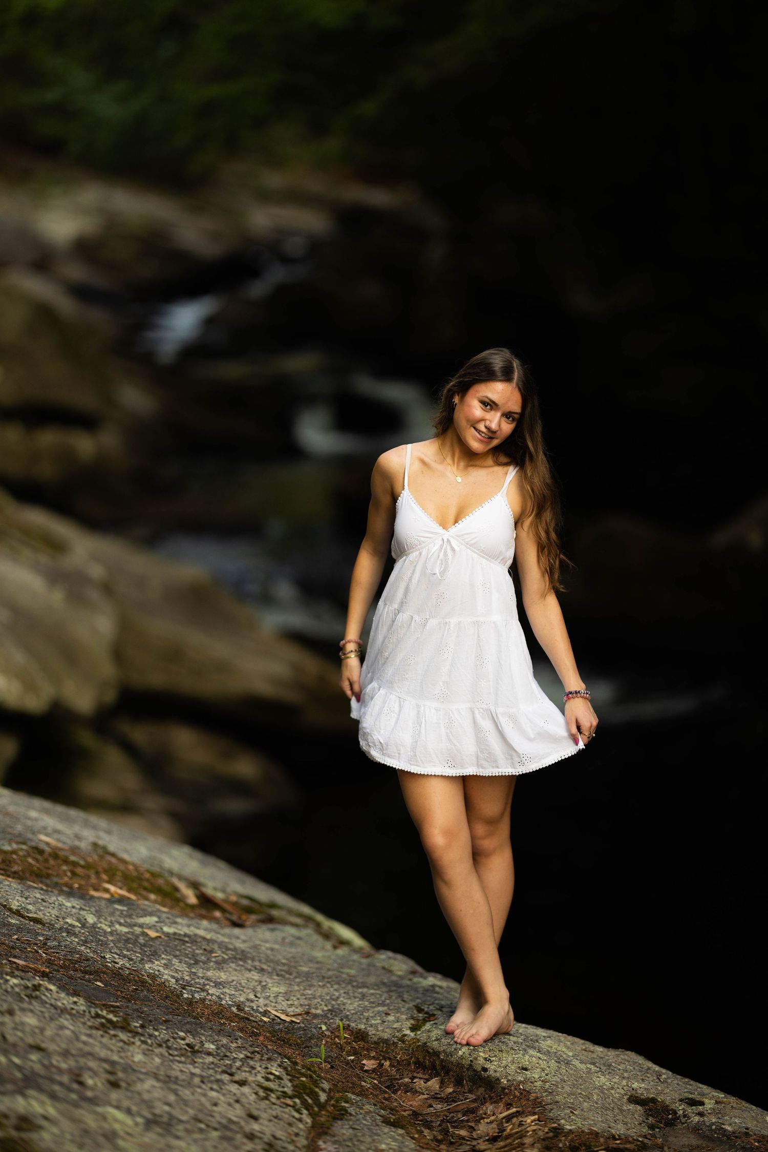 Young woman in white sundress standing barefoot on rocks near a waterfall in a lush forest setting.