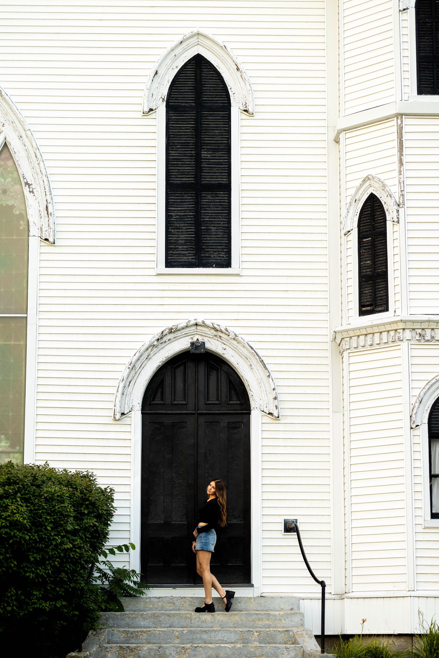 Young woman in black top and denim shorts standing on steps of a white wooden church with Gothic arched windows.