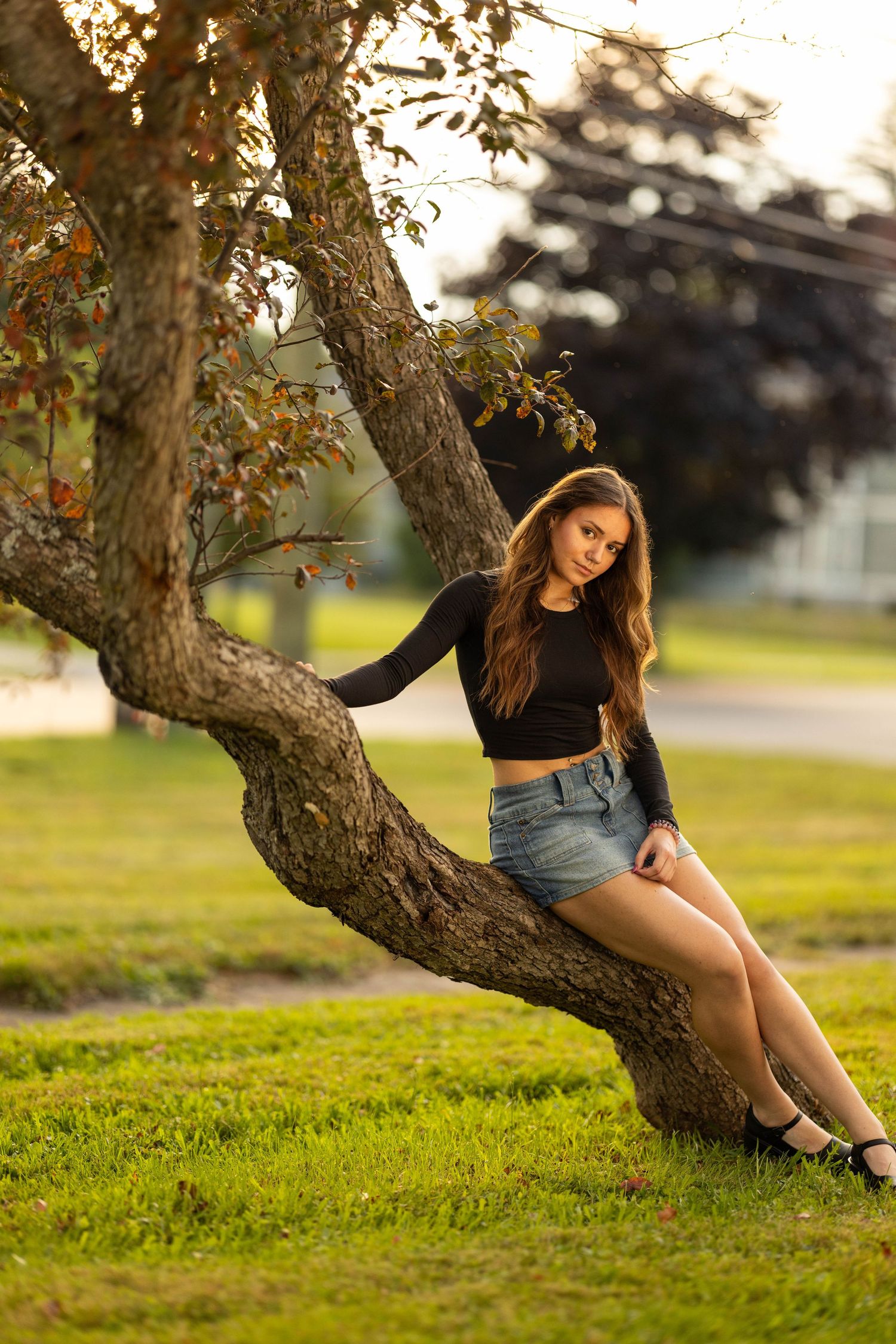 Young woman in black crop top and denim shorts sitting on a curved tree trunk in a sunny green park.