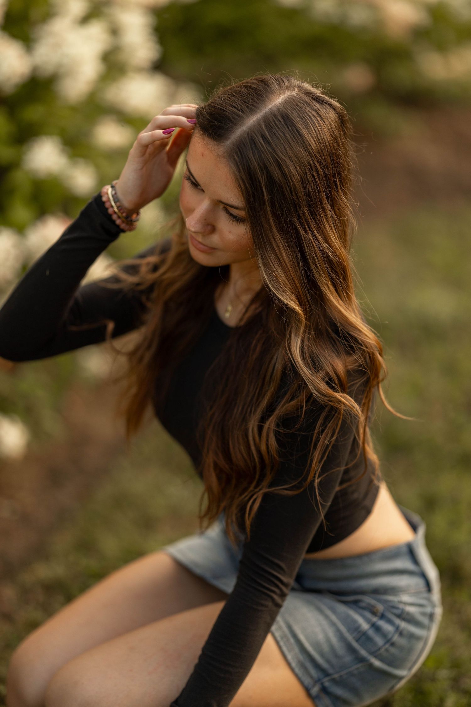 Young woman with long wavy brunette hair in black crop top and denim shorts poses outdoors near blooming white flowers.