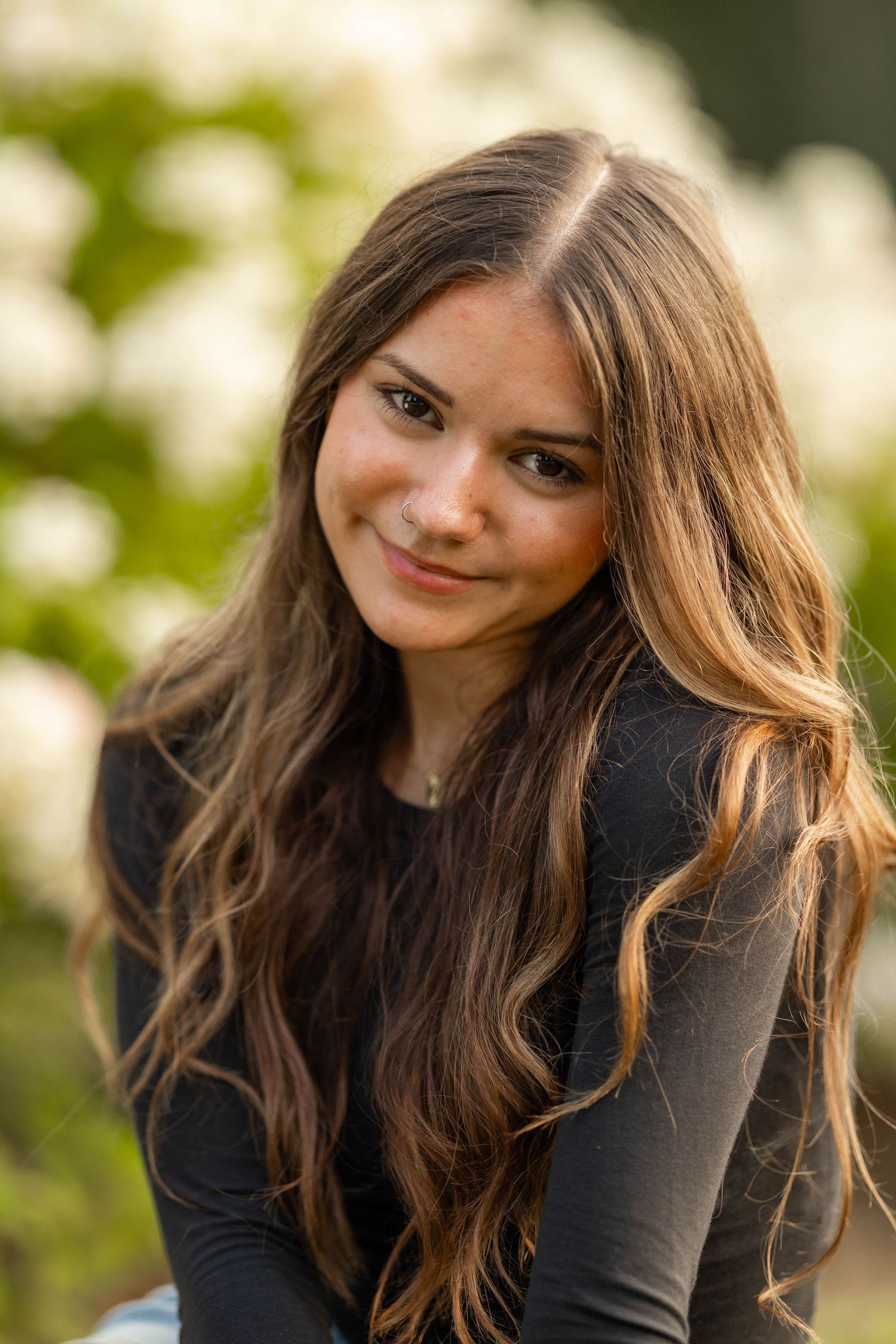 Young woman with long brown wavy hair smiling softly outdoors with white flowers in background.