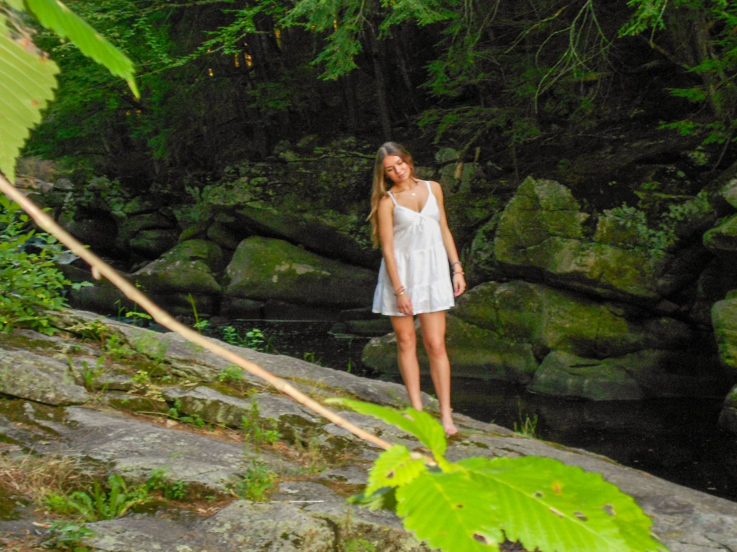 Young woman in white dress standing on mossy rocks near a forest waterfall surrounded by lush green foliage.