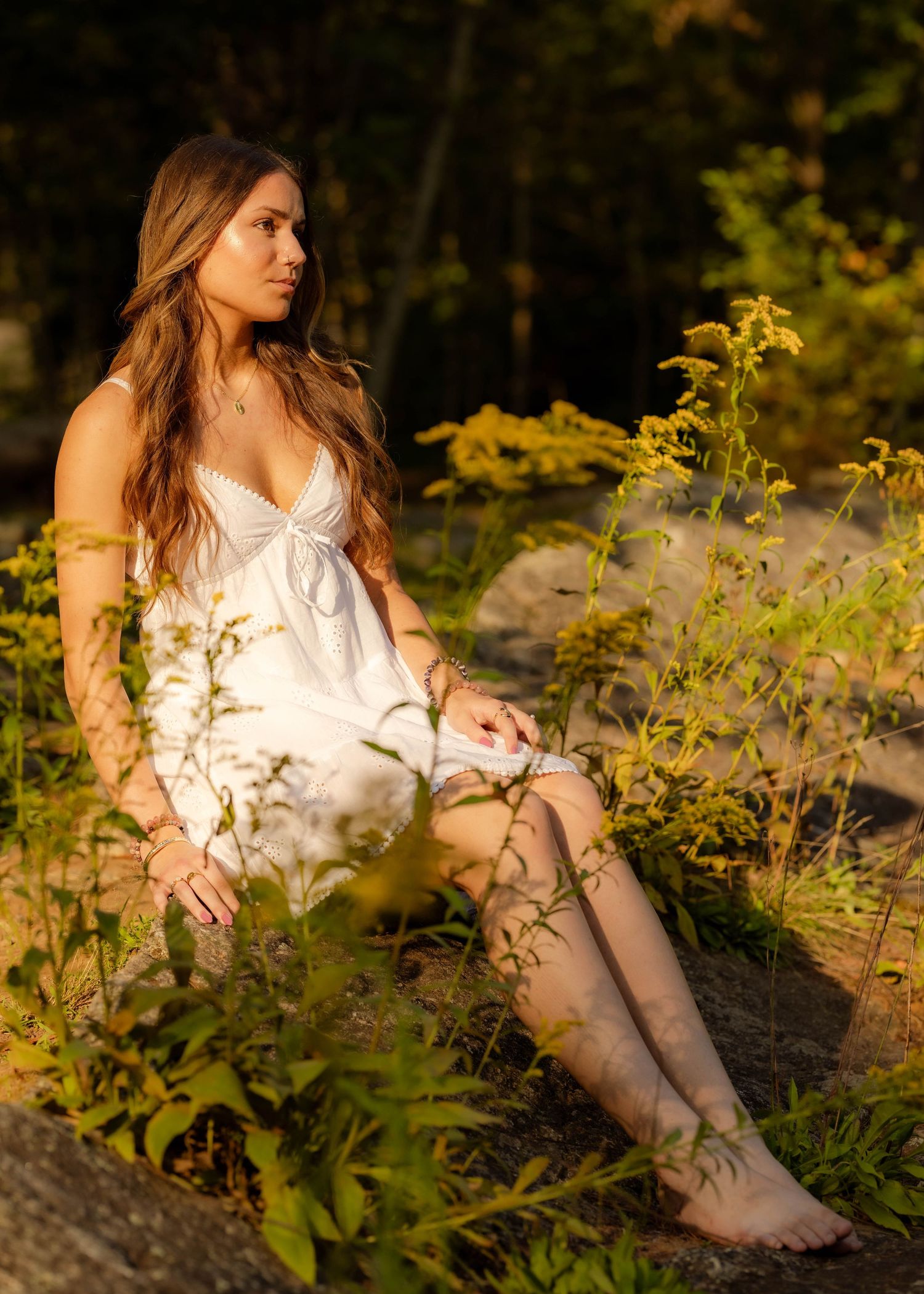 Young woman in white dress sitting barefoot among wildflowers in golden forest light.