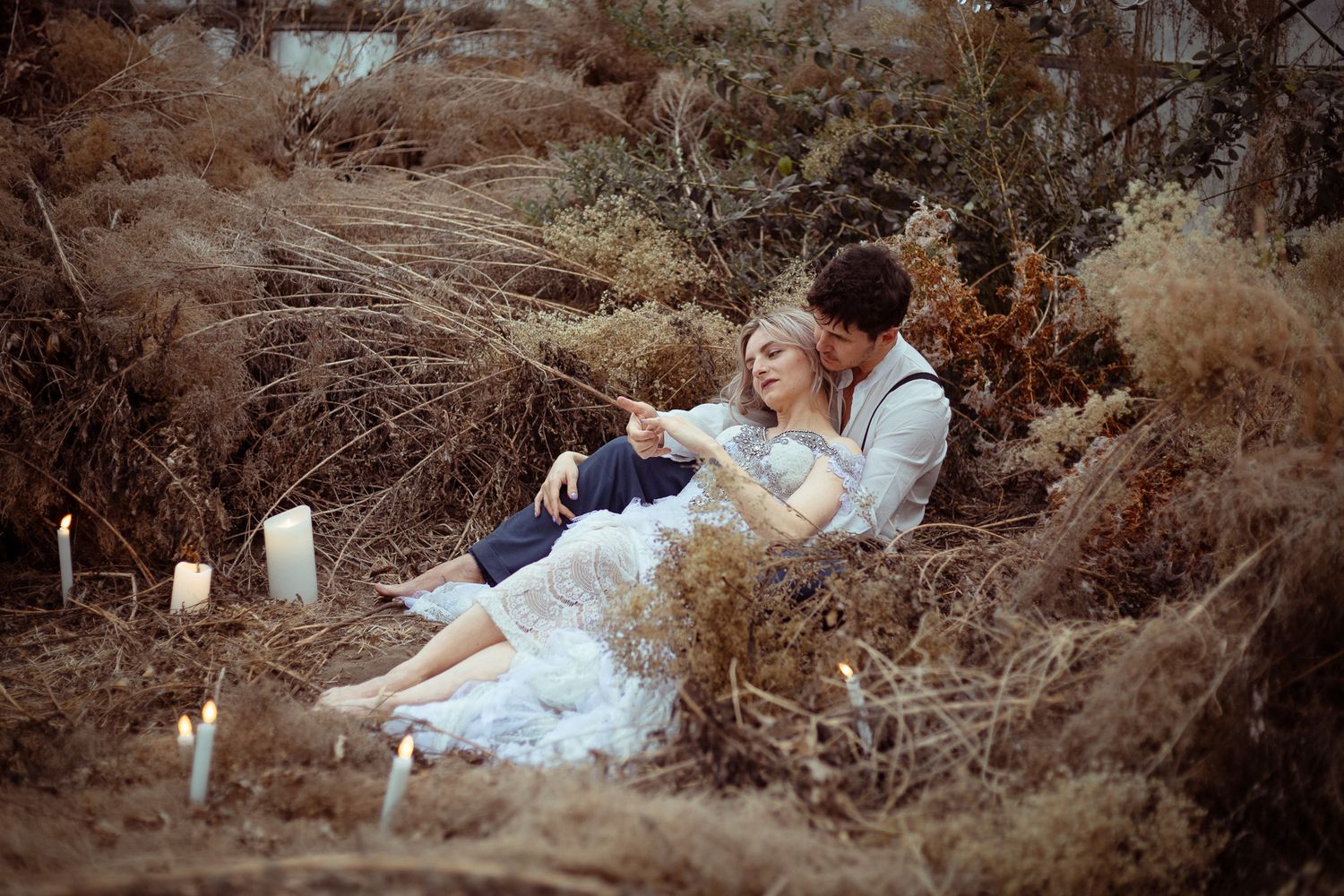 A romantic couple sits together among tall dried grass and candles in a dreamy outdoor setting.