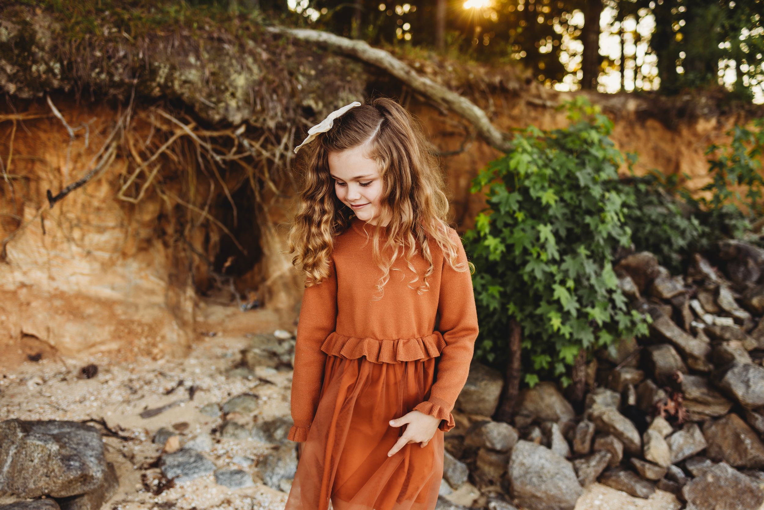 Woman in rust-colored dress stands against rocky coastal backdrop.