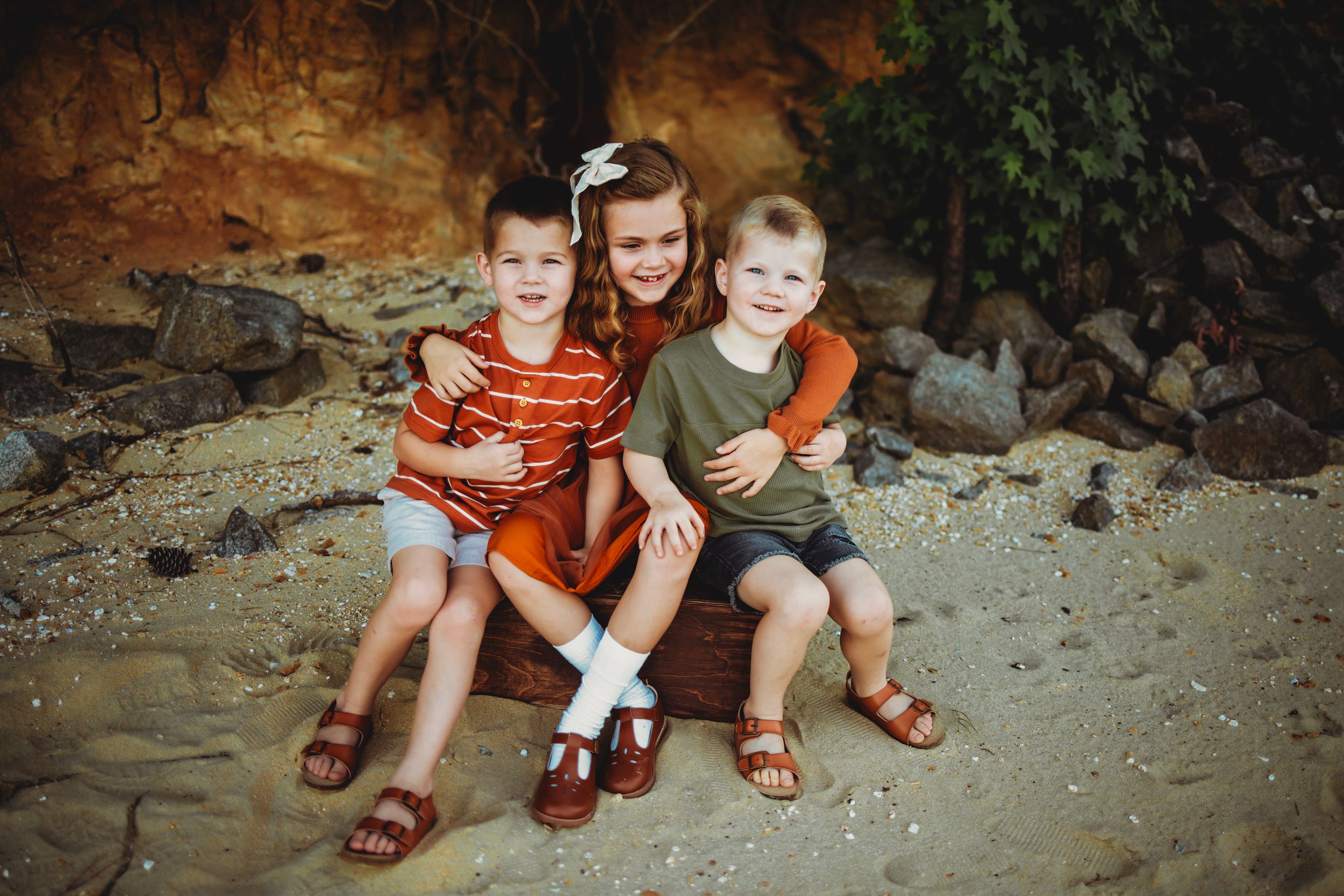 Three children sit together on sandy ground against rocky backdrop.