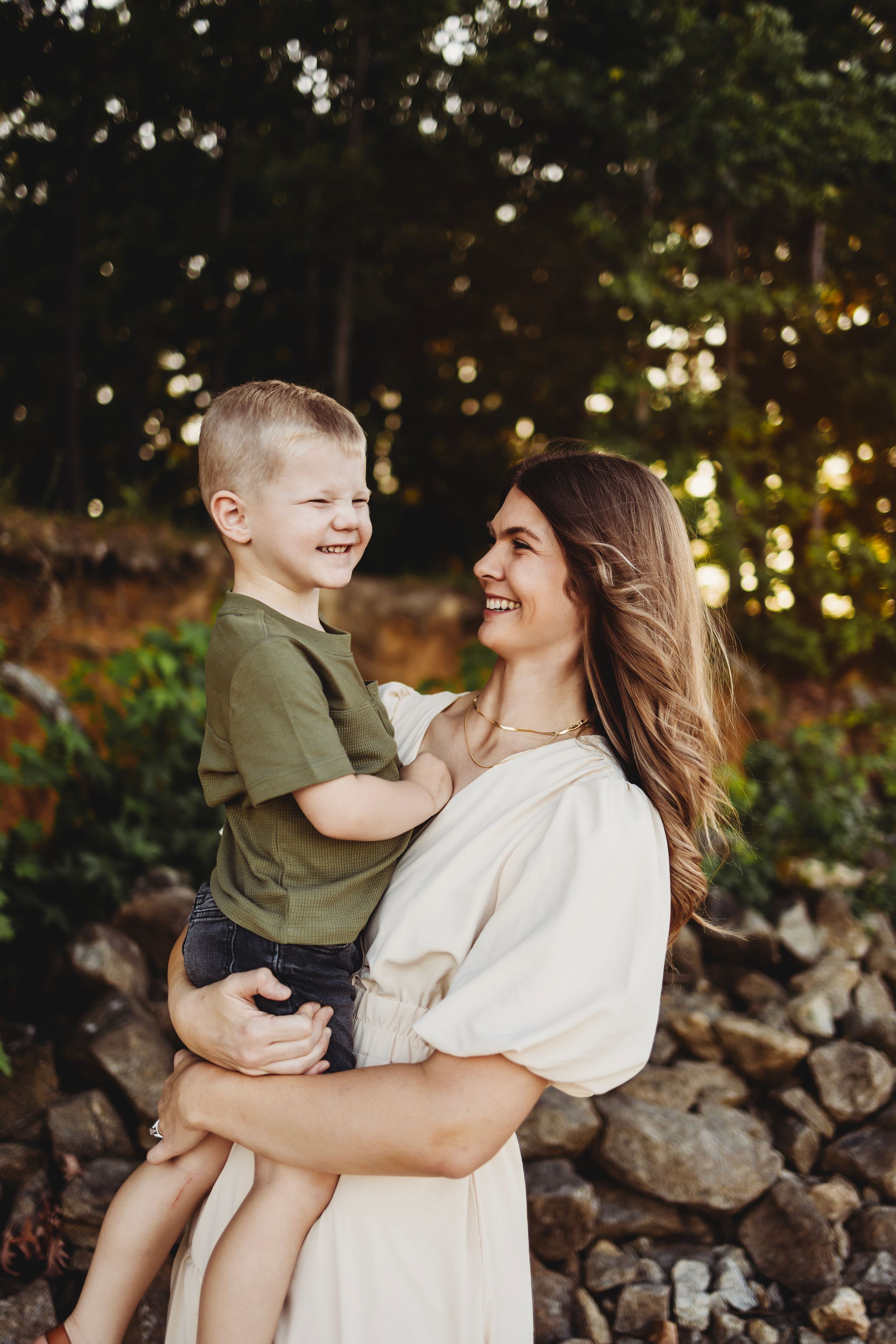 Joyful moment captured between parent and child against stone wall with sunlit trees.
