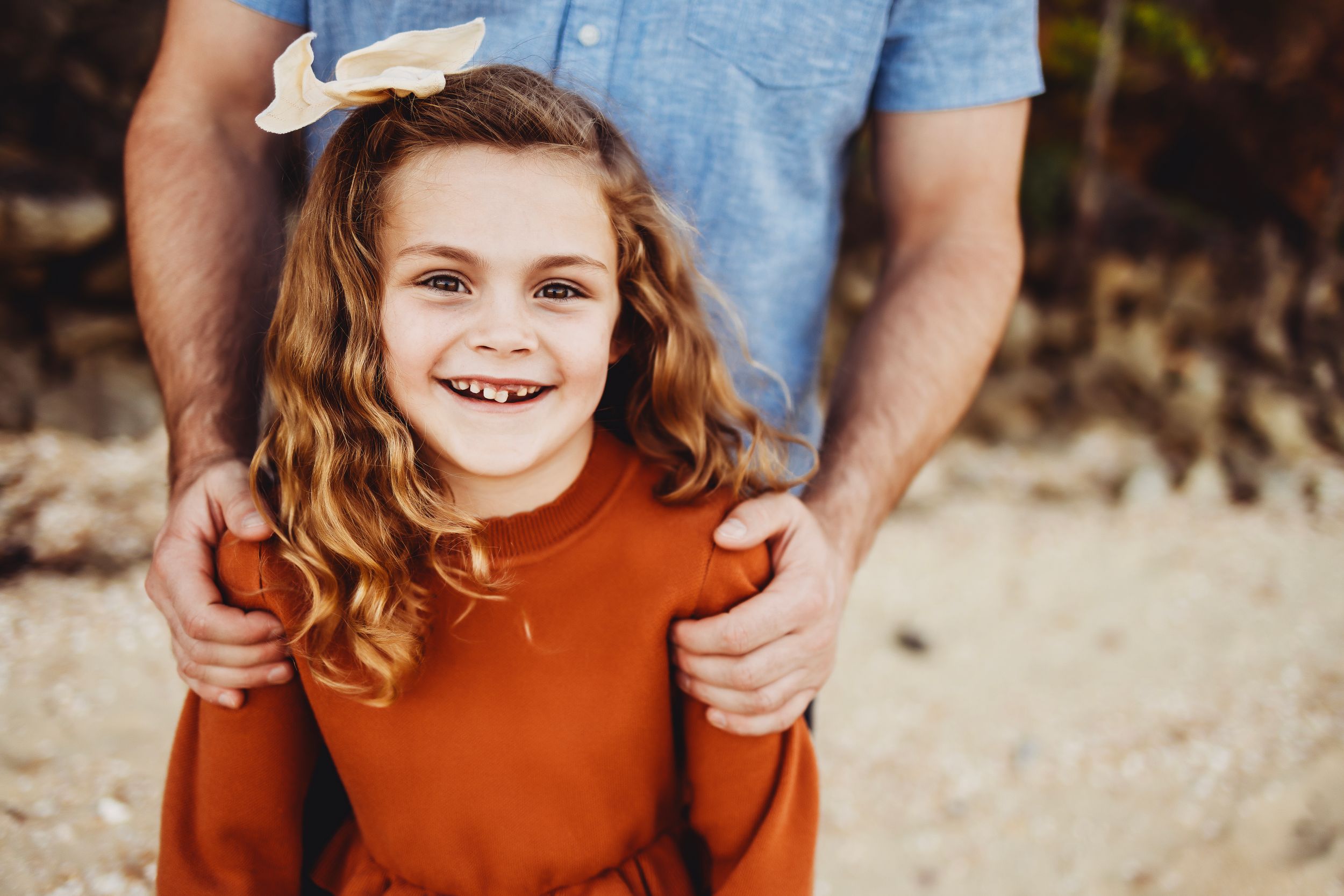 Smiling child with curly hair wearing a rust-colored sweater outdoors.