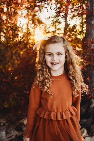 Child in rust-colored ruffle dress poses against autumn foliage at sunset.